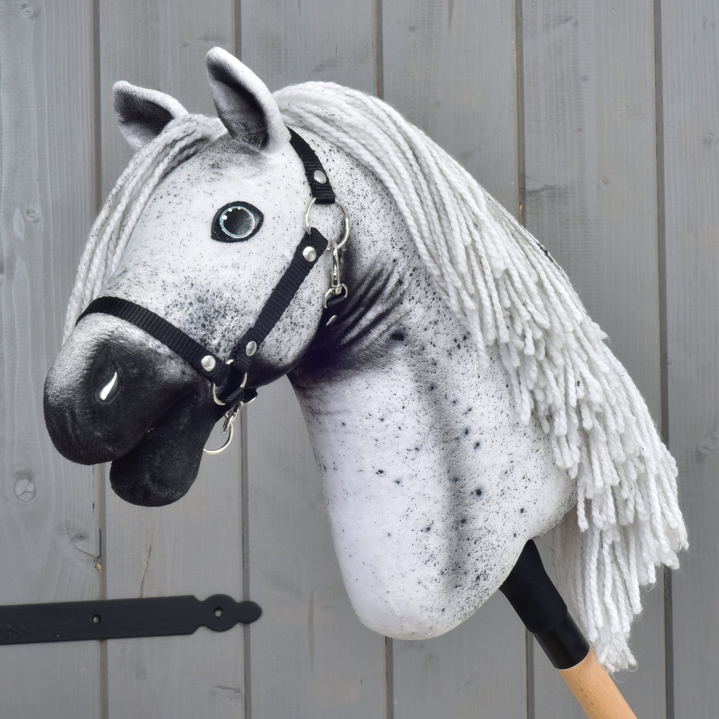 Hobby Horse Naivy avec licol noir - taille S de Hobby Horse LarDen, avec un design Appaloosa gris et blanc, un licol noir et une crinière en fil, monté sur un bâton en bois et placé devant un mur en bois gris.