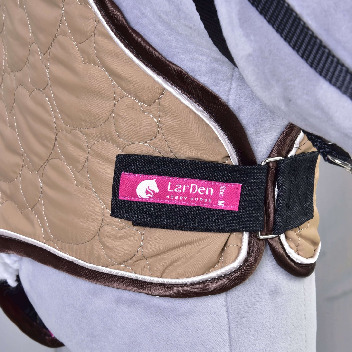 Nahaufnahme eines hellbraun gesteppten Halsteil Bonanza von Hobby Horse LarDen mit pinkem Label und schwarzem Riemen, gezeigt an einem hellgrauen Hobby Horse. Sichere Verschlüsse halten es beim Spielen perfekt an Ort und Stelle.