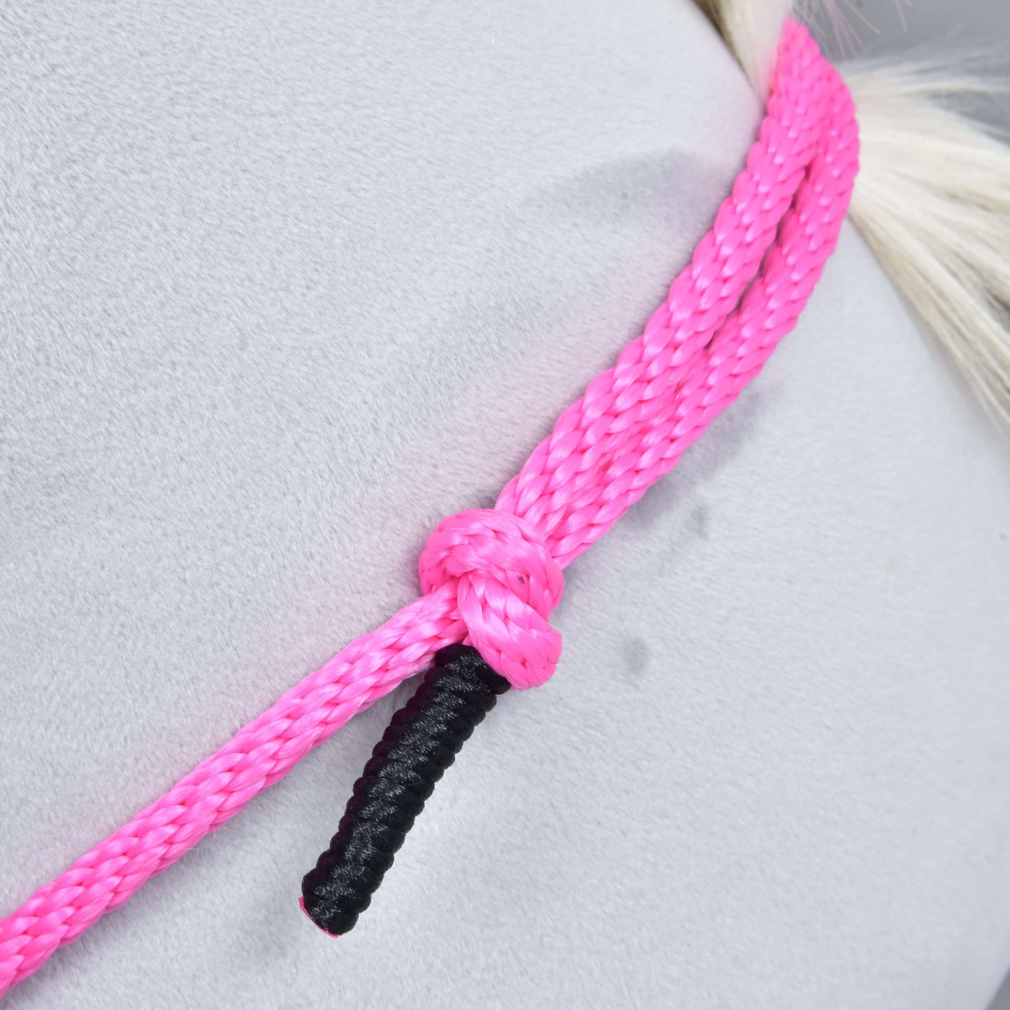 Nahaufnahme von Halsring Pink Festive von Hobby Horse LarDen – ein leuchtend pinkes handgefertigtes Cordeo-Seil mit einem schwarz endenden Strang, um eine hellgraue Oberfläche geknotet. Ideal für gebissloses Reiten; der Hintergrund ist glatt mit schwachen Spuren.