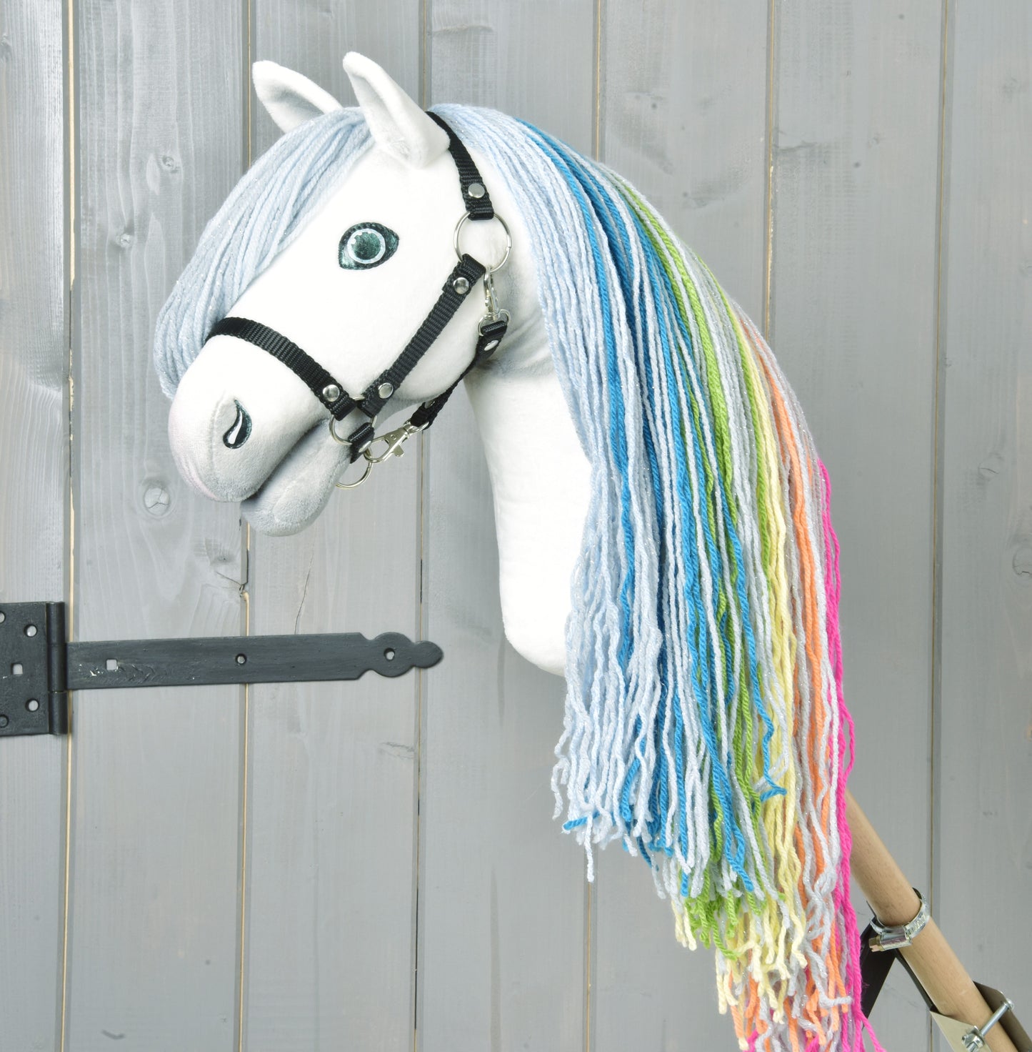 Hobby Horse Rainbow New de Hobby Horse LarDen avec visage blanc fait main, yeux bleus, bride noire et crinière arc-en-ciel en fil bleu, vert, jaune et rose, sur fond en bois gris clair pour une touche féerique.