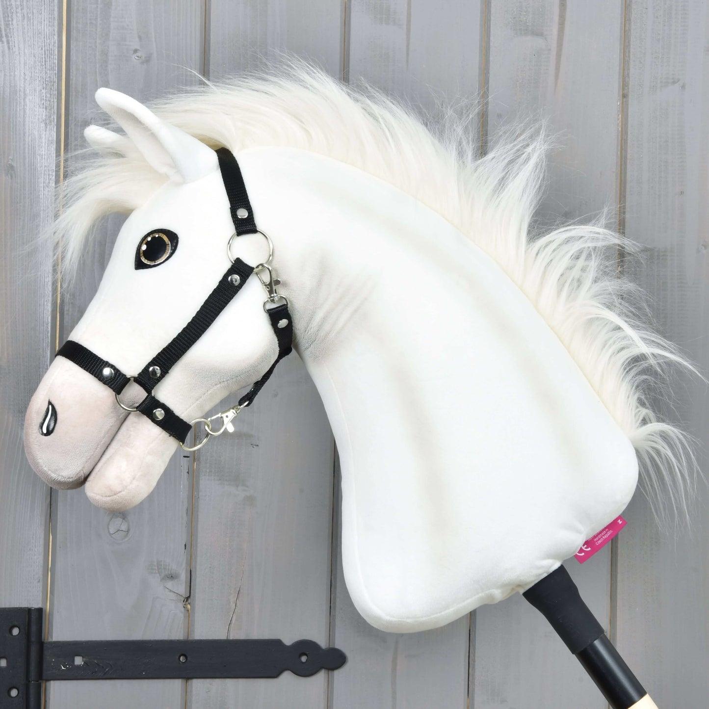 Hobby Horse LarDen Hobby Horse Nexus mit Schwarzem Halfter mit einem weißen Sportpferd aus Plüsch mit flauschiger Mähne, großem Knopfauge und schwarzem Halfter – ideal für jeden Fan. Fotografiert vor einer grauen Holzwand.