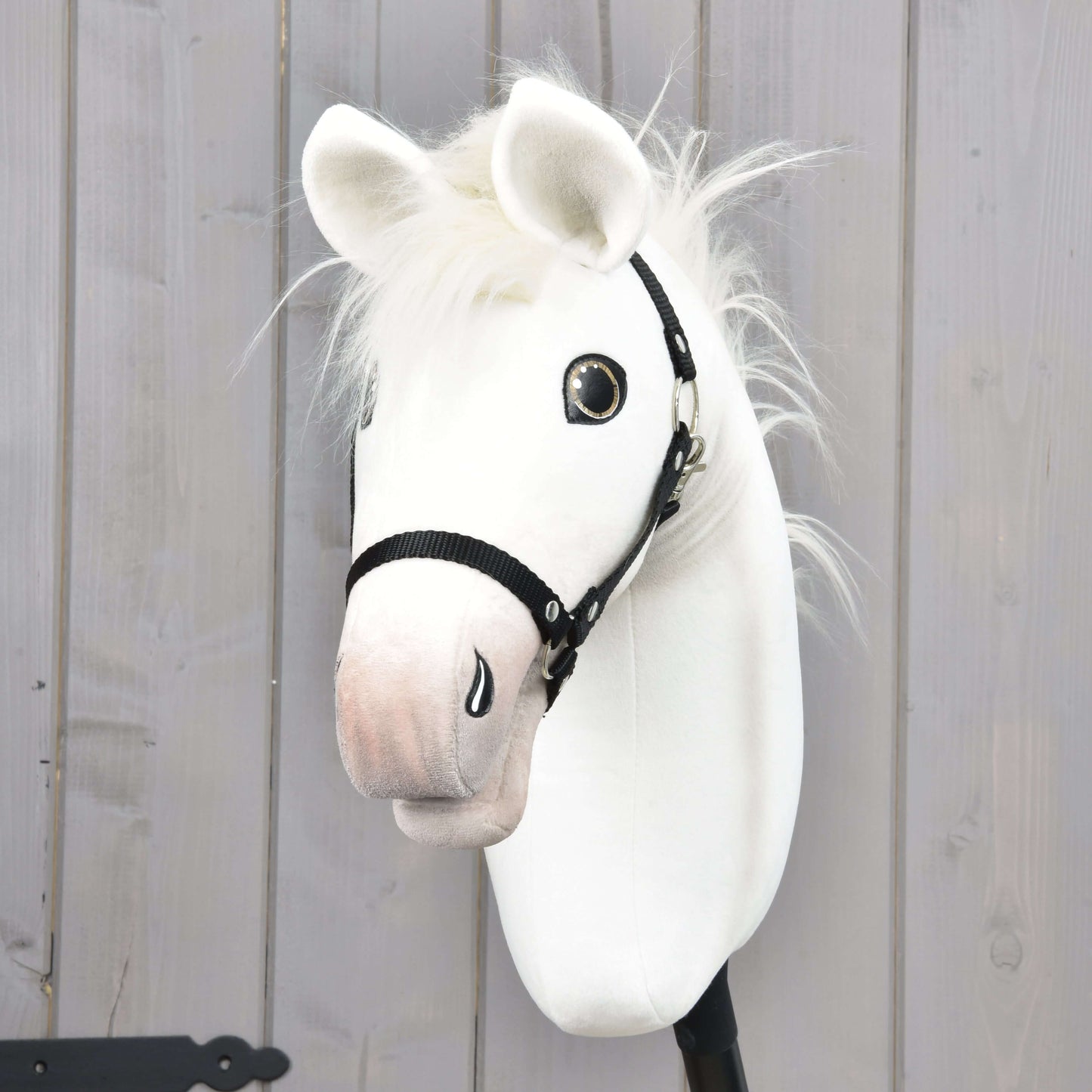 Hobby Horse Nexus mit Schwarzem Halfter von Hobby Horse LarDen, ein weißes Stoff-Hobby Horse mit weicher Mähne und glänzenden Augen, vor einem Hintergrund aus hellen Holzpaneelen.