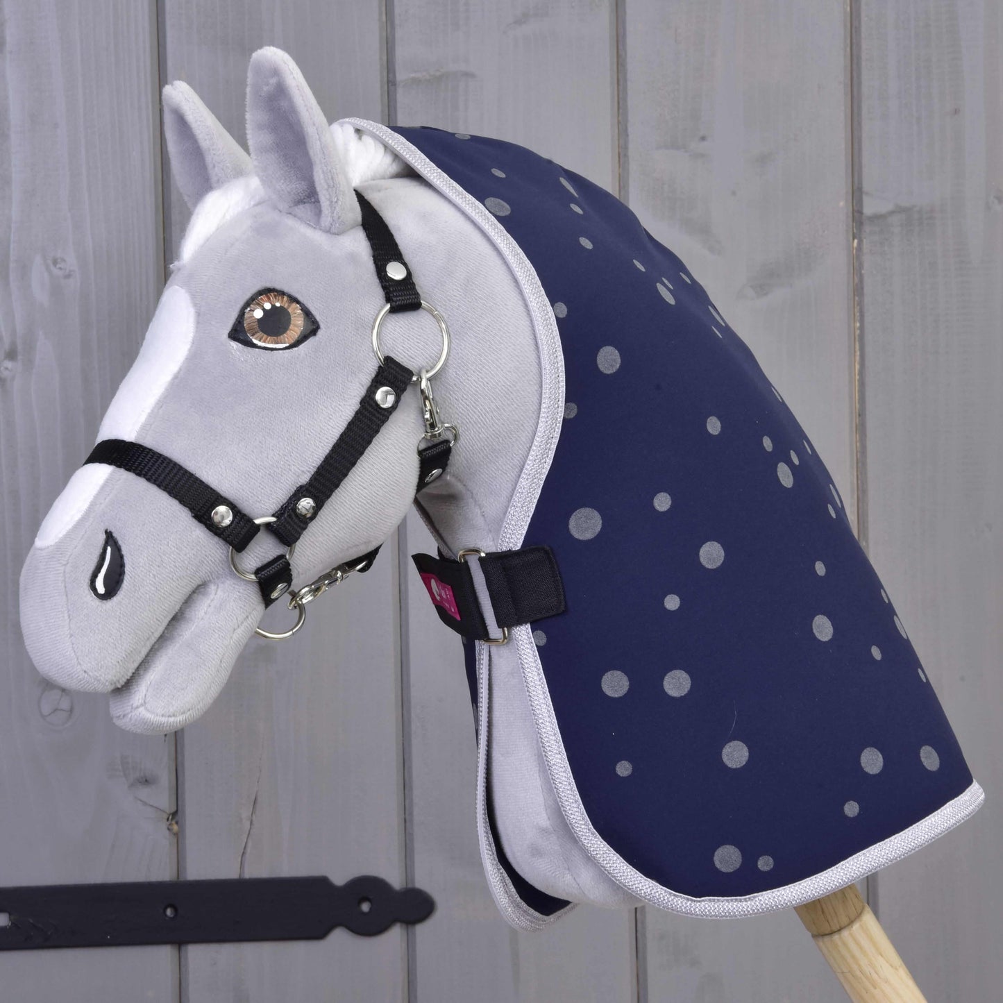 Hobby Horse en peluche gris de Hobby Horse LarDen avec bride noire et Couverture d'écurie Ciel de Nuit, monté sur un bâton en bois devant un mur en bois clair.