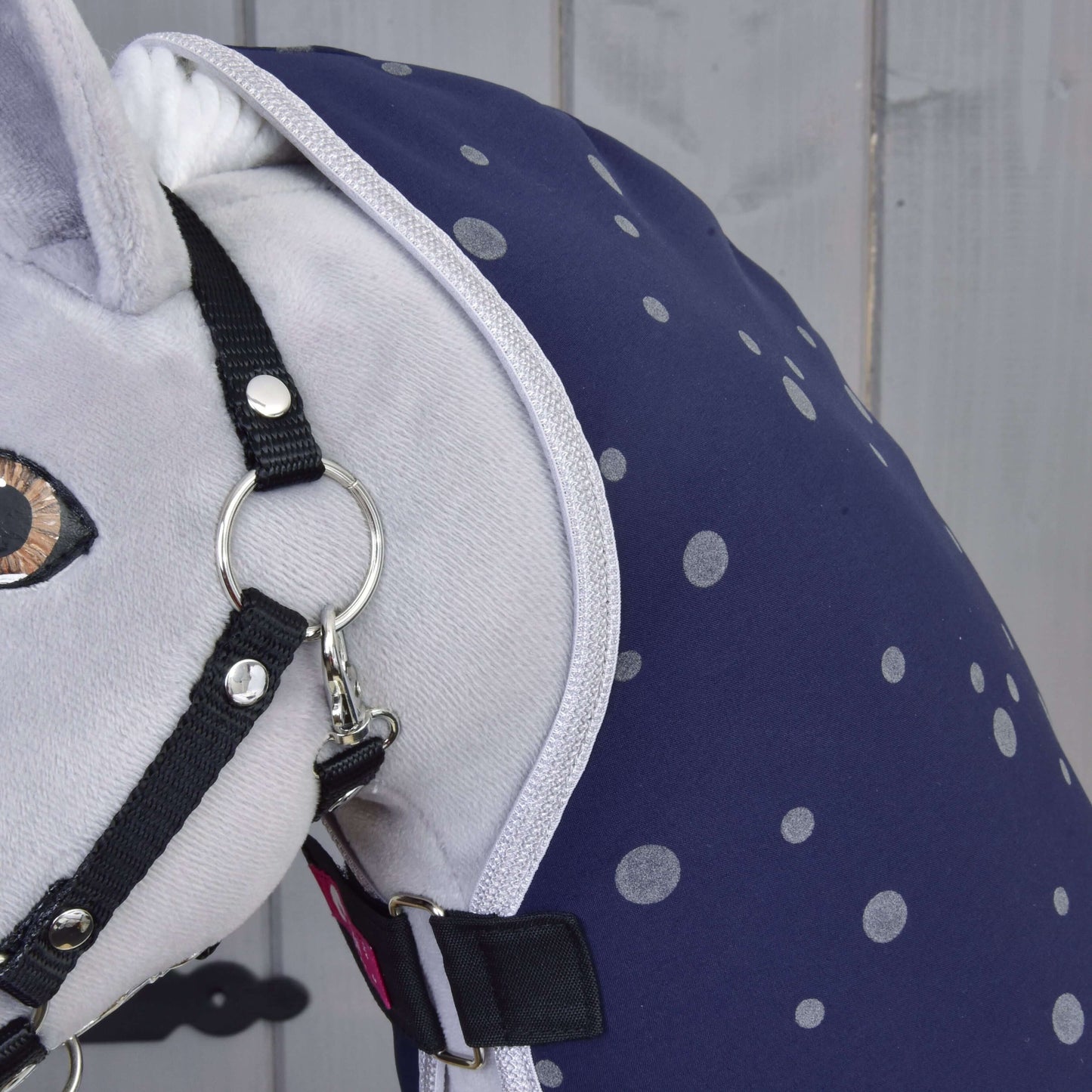 Gros plan sur un cheval en peluche portant la Couverture d'écurie Ciel de Nuit de Hobby Horse LarDen, bleu marine à pois gris clair, avec un licol noir, sur fond en bois gris clair.