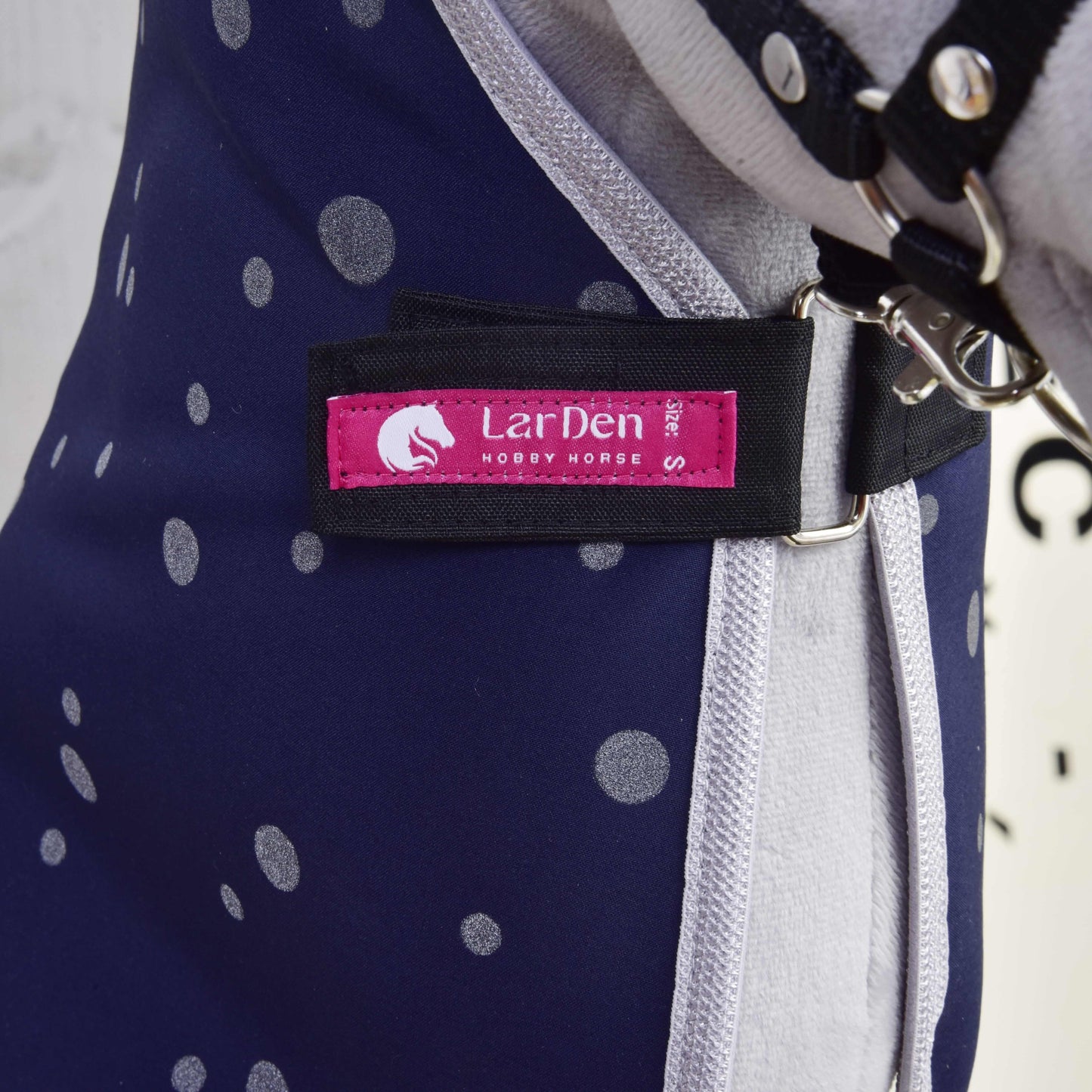Gros plan sur la Couverture d'écurie Ciel de Nuit de Hobby Horse LarDen, montrant un tissu bleu foncé à pois blancs, une étiquette de marque rose et noire, et un clip métallique argenté sur la sangle.