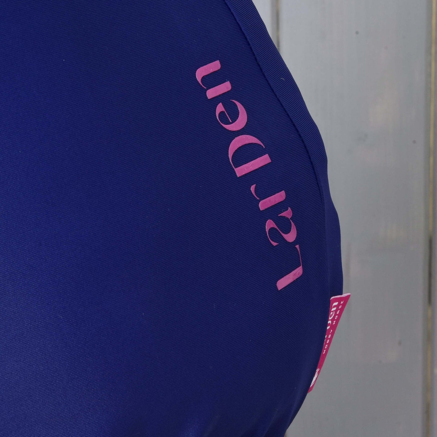 Gros plan sur la Combinaison de protection Hobby Horse LarDen en lycra bleu foncé de Hobby Horse LarDen, avec texte rose « LarDen » et recouvrant partiellement une surface en bois gris clair.