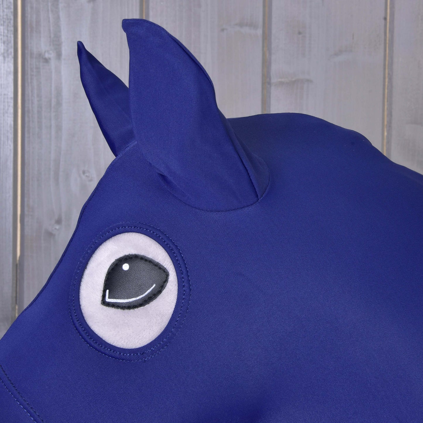 Gros plan sur la Combinaison de protection Hobby Horse LarDen en lycra bleu, conçue pour les Hobby Horses, avec grand œil de style cartoon et oreille pointue, présentée devant un fond en panneaux de bois clair.