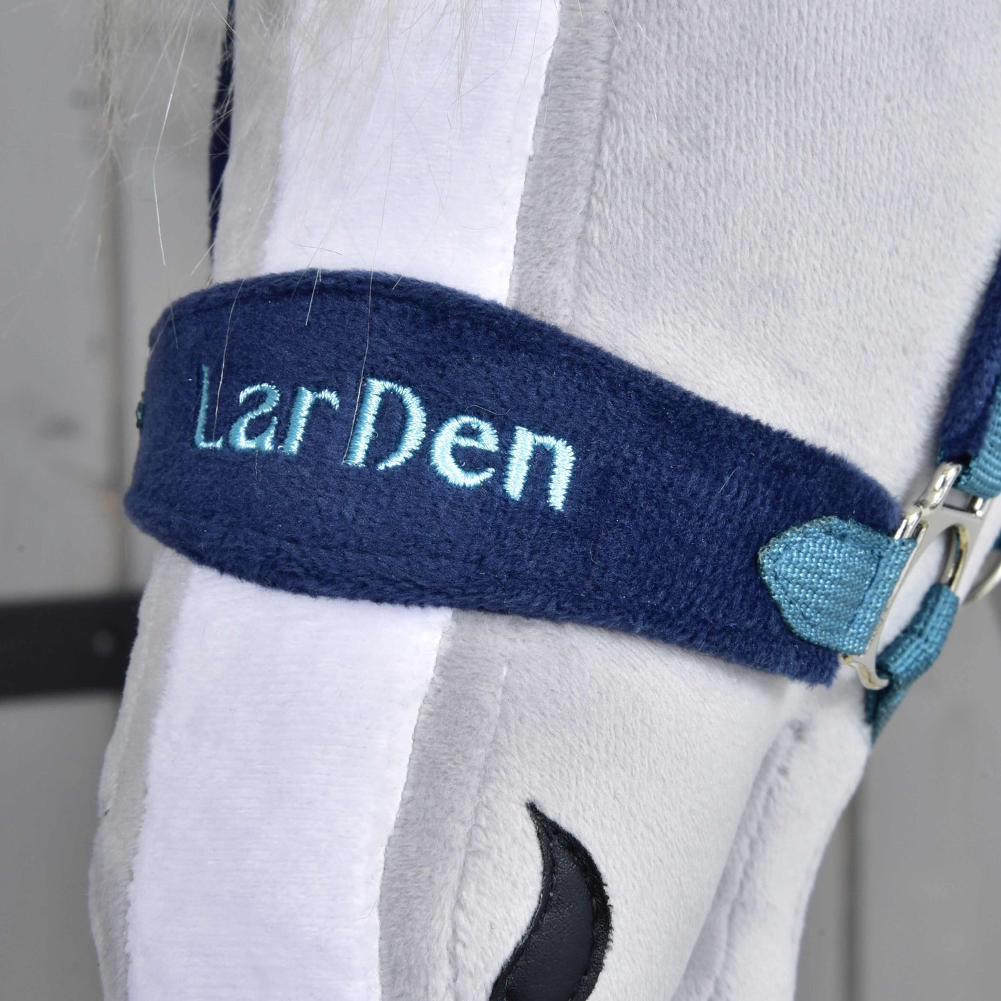 Gros plan sur une tête de cheval en peluche blanche portant le Licol en fourrure LarDen bleu de Hobby Horse LarDen, brodé d'un texte bleu clair, un ajout élégant à toute collection de licols pour Hobby Horse.