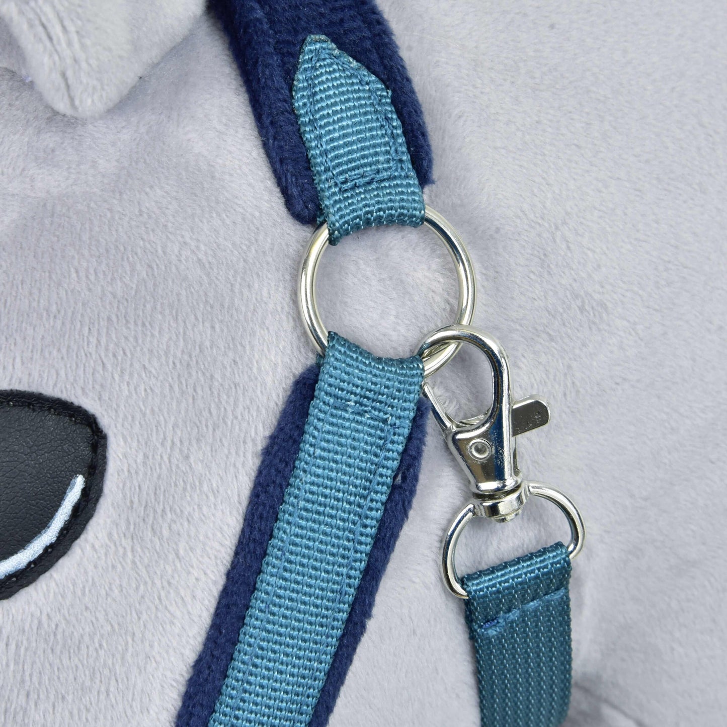 Gros plan sur le fermoir et l’anneau métalliques reliant deux sangles bleues en nylon sur une fourrure douce gris clair, avec un détail d’œil brodé noir et blanc du Licol en fourrure LarDen bleu de Hobby Horse LarDen.