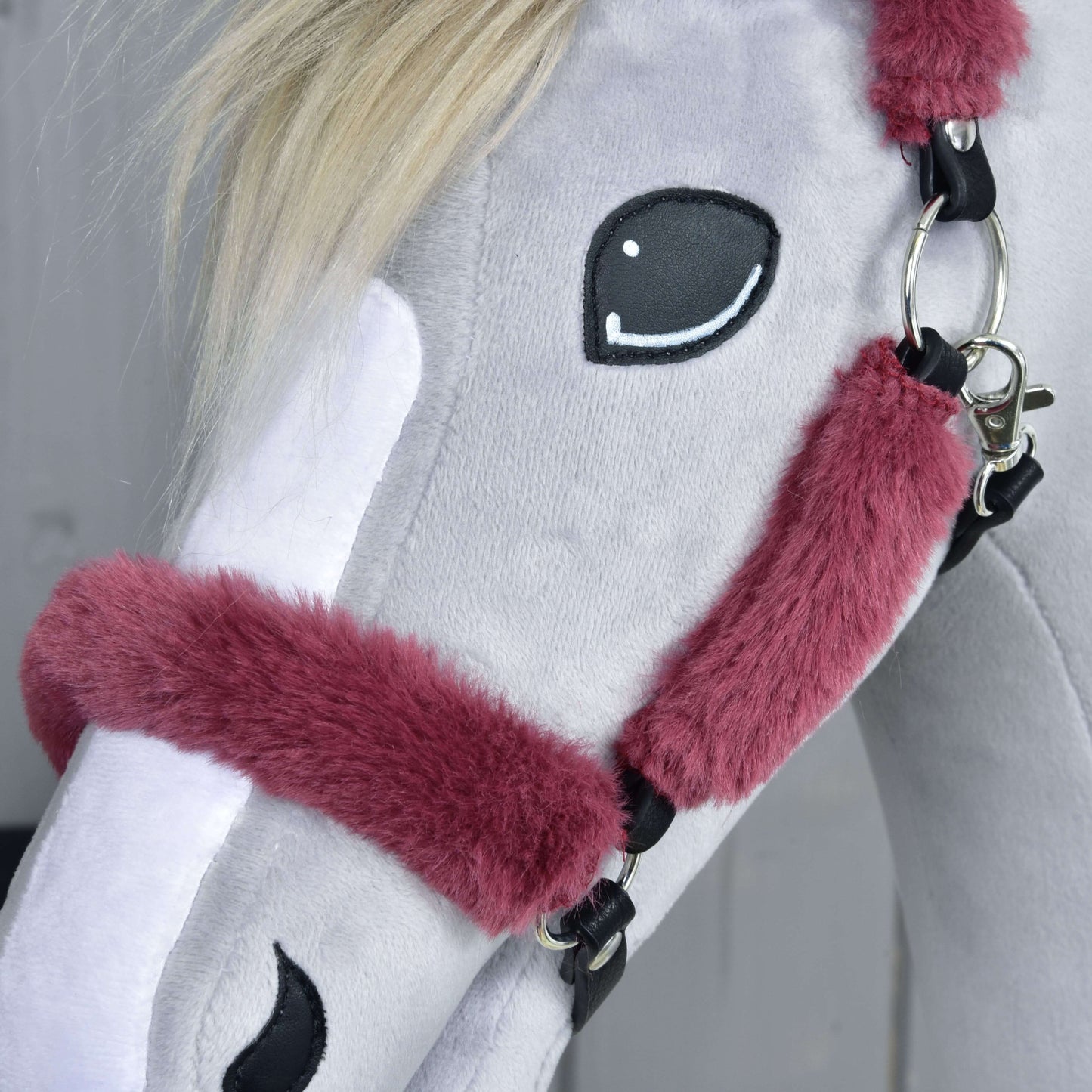 Gros plan sur une peluche grise Hobby Horse LarDen, Licol en fourrure Teddy bordeaux, avec licol en polaire bordeaux, crinière blanche, yeux brodés et marque blanche sur le visage. Arrière-plan flou.