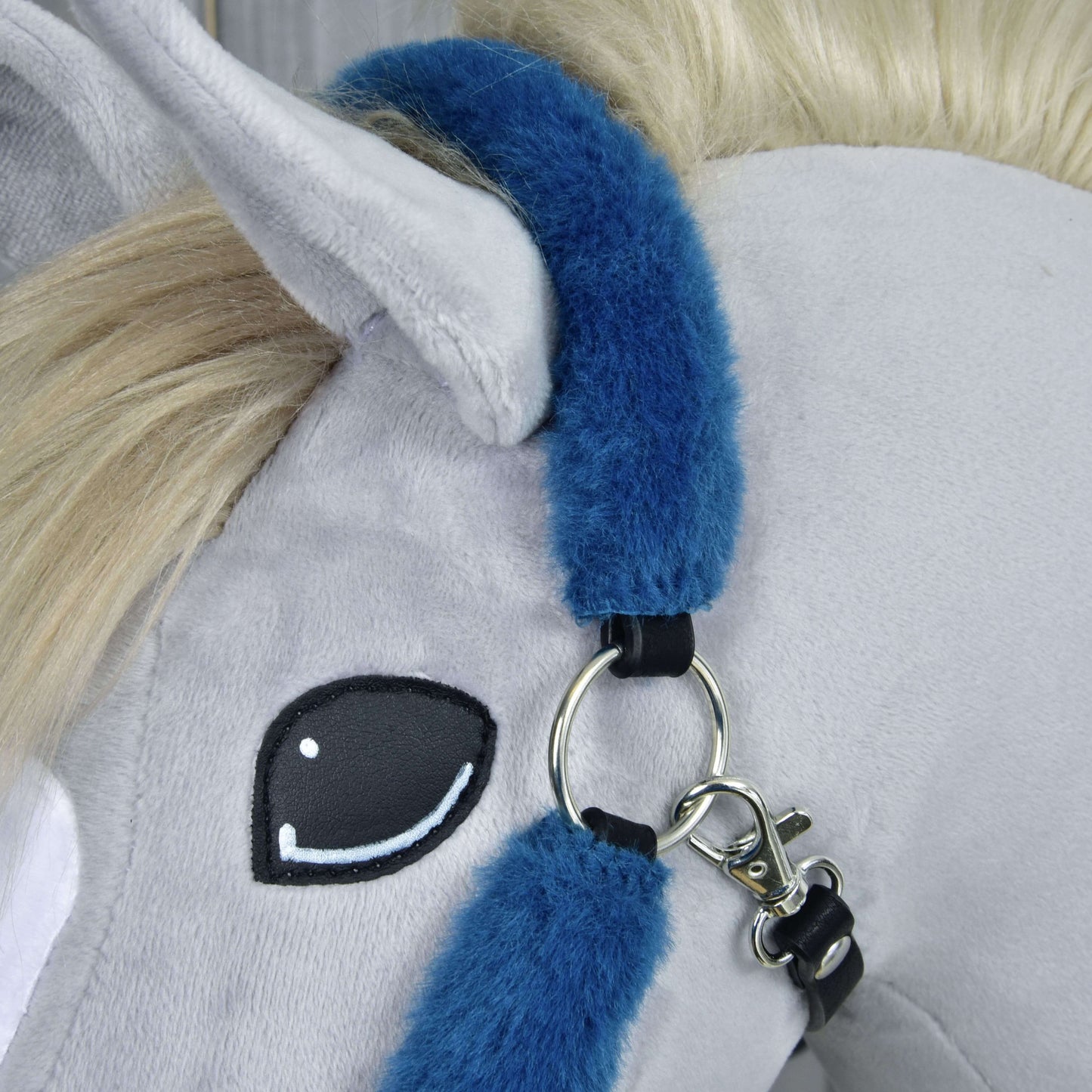 Gros plan sur une tête de cheval en peluche grise avec crinière crème, œil brodé noir et blanc, portant le Licol en fourrure Teddy pétrole de Hobby Horse LarDen en bleu avec anneaux et fermoir métalliques.