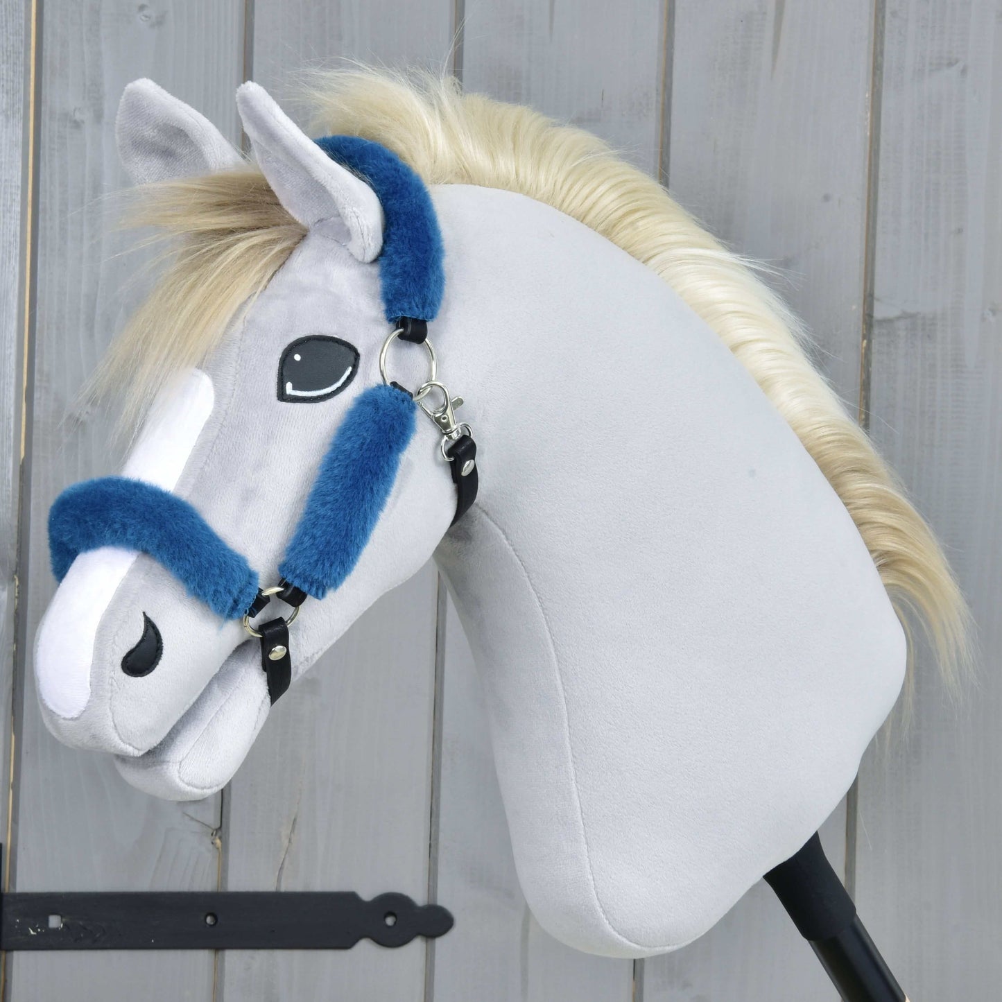 Hobby Horse LarDen Licol en fourrure Teddy pétrole, un Hobby Horse en peluche avec tissu gris clair, museau blanc, crinière blonde et licol bleu en similicuir sur un bâton noir, présenté devant un fond en bois.