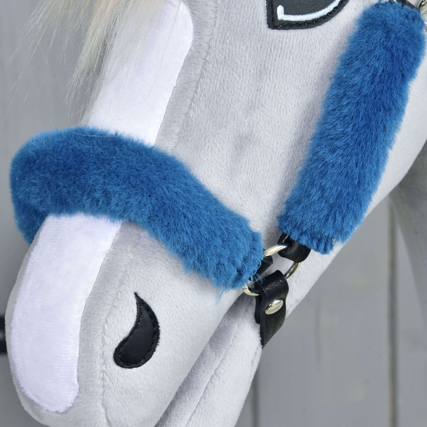 Gros plan sur une tête de cheval en peluche gris clair portant le Licol en fourrure Teddy pétrole de Hobby Horse LarDen, avec licol bleu, crinière blanche et détails d’œil et de narine cousus en noir, sur un fond neutre doucement flou.