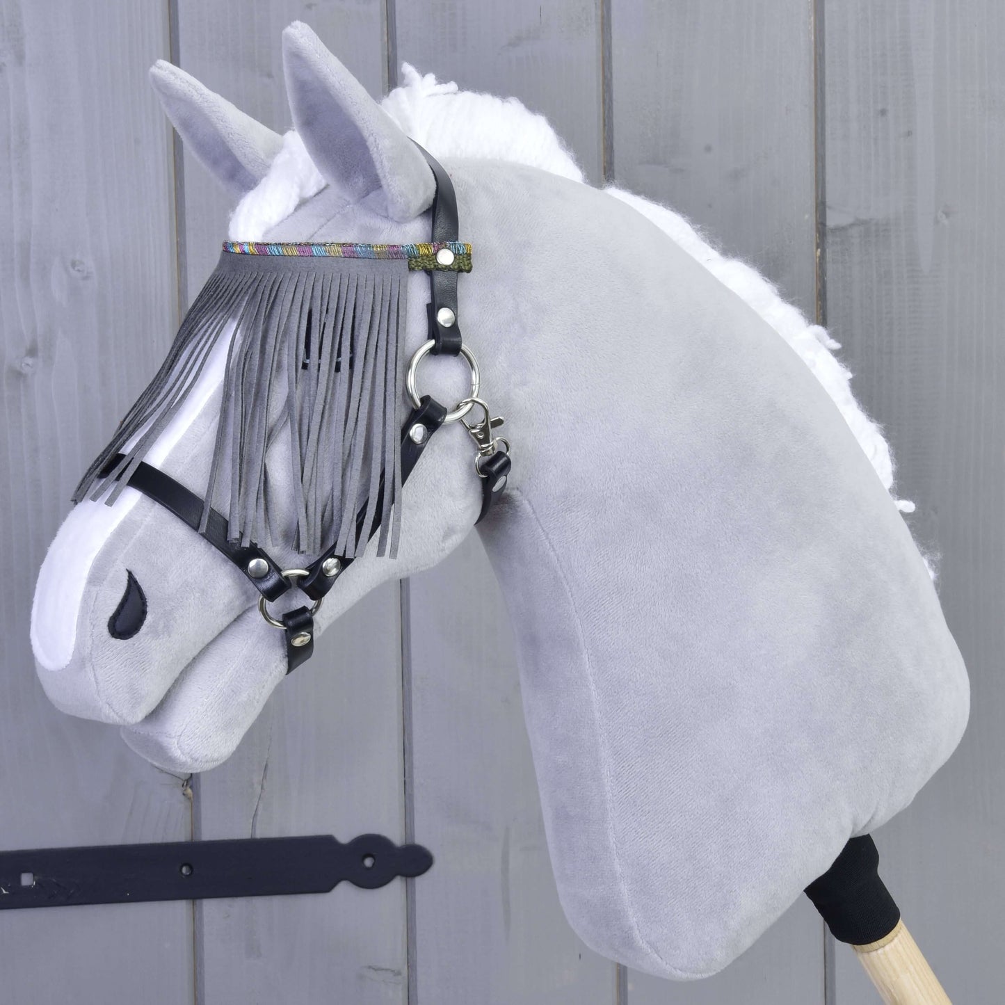 A plush gray hobby horse head with a white mane, wearing the Hobby Horse LarDen halter in black with grey fringes and a gray browband, mounted on a wooden stick, shown against a wooden wall.