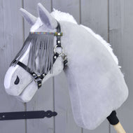 A plush gray hobby horse head with a white mane, wearing the Hobby Horse LarDen halter in black with grey fringes and a gray browband, mounted on a wooden stick, shown against a wooden wall.