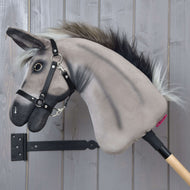 Hobby Horse LarDen „Esel Olin mit Schwarzem Halfter“ mit grauem Plüsch-Eselkopf, dunkler Mähne, schwarzem Halfter für Hobby Horse, Zügeln und silbernem Ring nahe dem Maul auf einem Holzstab, vor grauer Wand.
