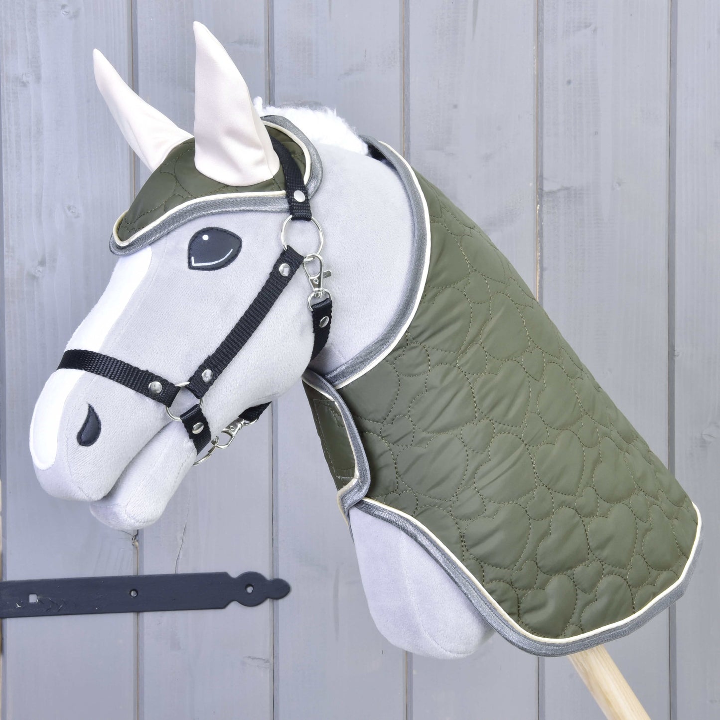 Das Set Olivetti von Hobby Horse LarDen zeigt ein graues Hobby Horse mit grüner gesteppter Decke und Zaumzeug, schwarzem Halfter für Hobby Horse, weißen Ohren und stilvollen Accessoires vor einer hellgrauen Holzwand.