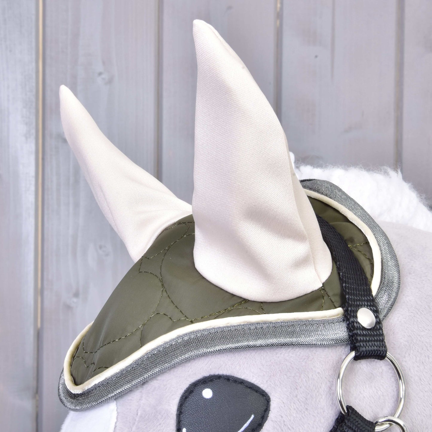 Nahaufnahme eines Plüschpferds mit dem Set Olivetti von Hobby Horse LarDen: eine grüne gesteppte Ohrenkappe mit zwei großen hellen Ohren, schwarzem Riemen und Metallring – einzigartiges Pferdezubehör auf Holzhintergrund.