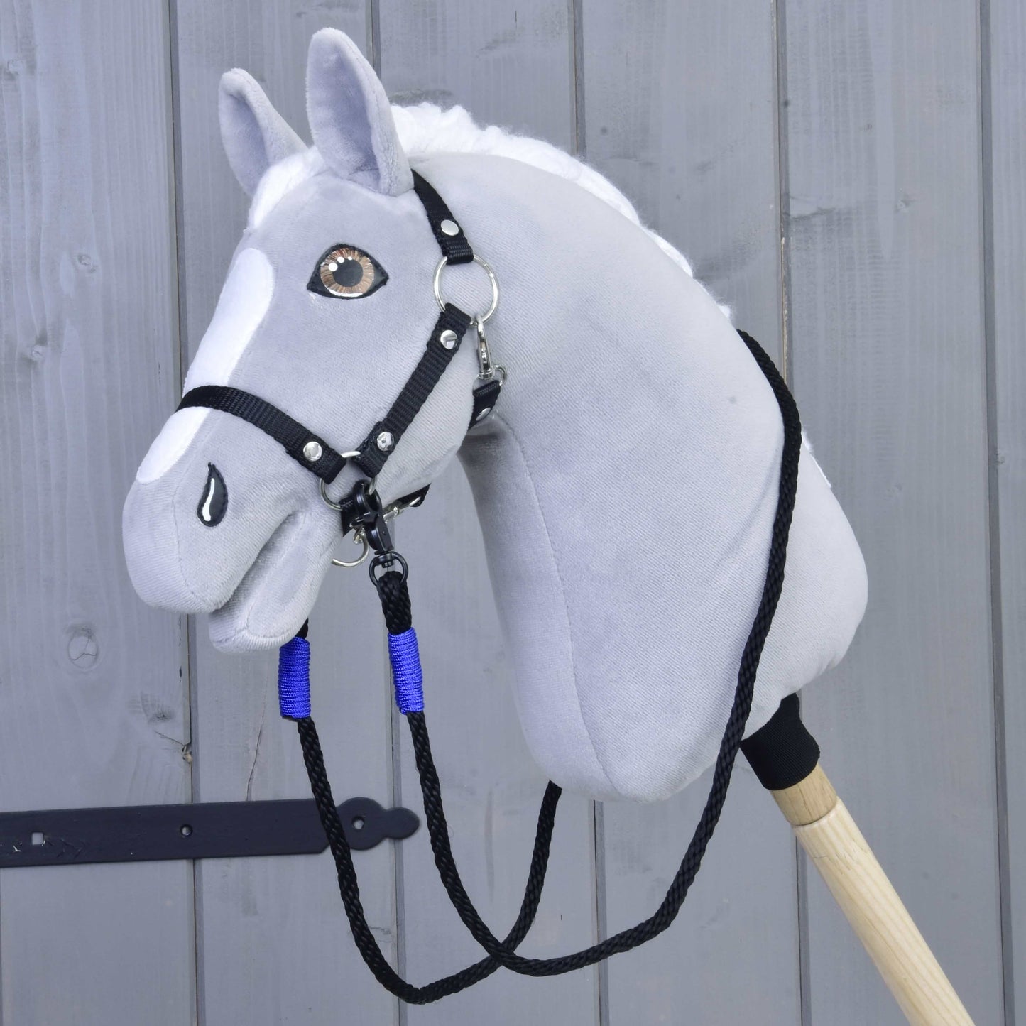 Hobby Horse LarDen Zügel Basic Blue an einem grauen Hobby Horse Kopf aus Plüsch mit schwarzem Zaumzeug für Hobby Horse und blauen Zügeln aus Polypropylenseil auf einem Holzstab vor einer hellgrauen Holzwand.