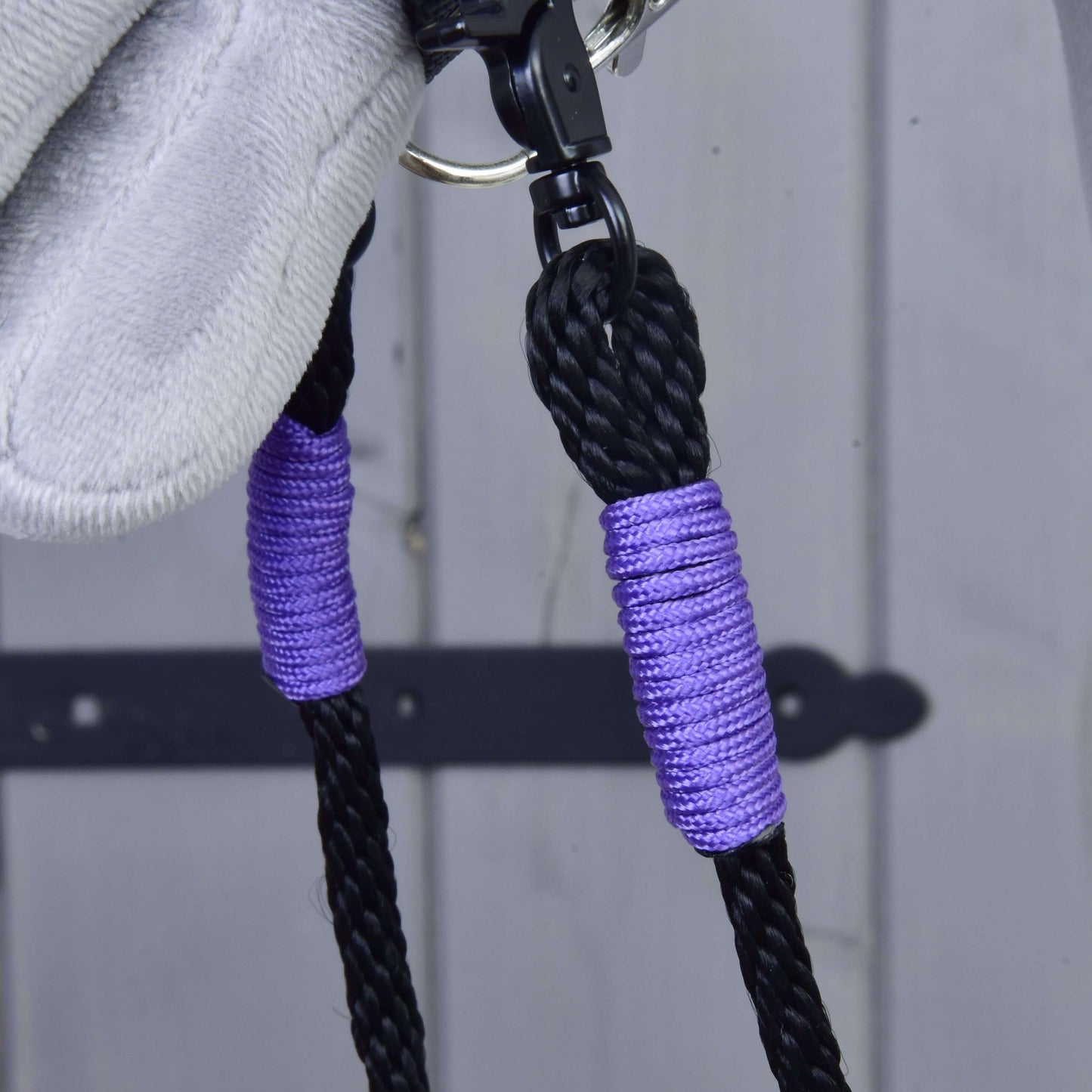 Nahaufnahme von Hobby Horse LarDen Zügel Basic Lila: schwarze handgefertigte Zügel aus Polypropylen mit lila Akzenten und silbernem Clip vor einer hellgrauen Holzoberfläche mit schwarzem Metallscharnier.