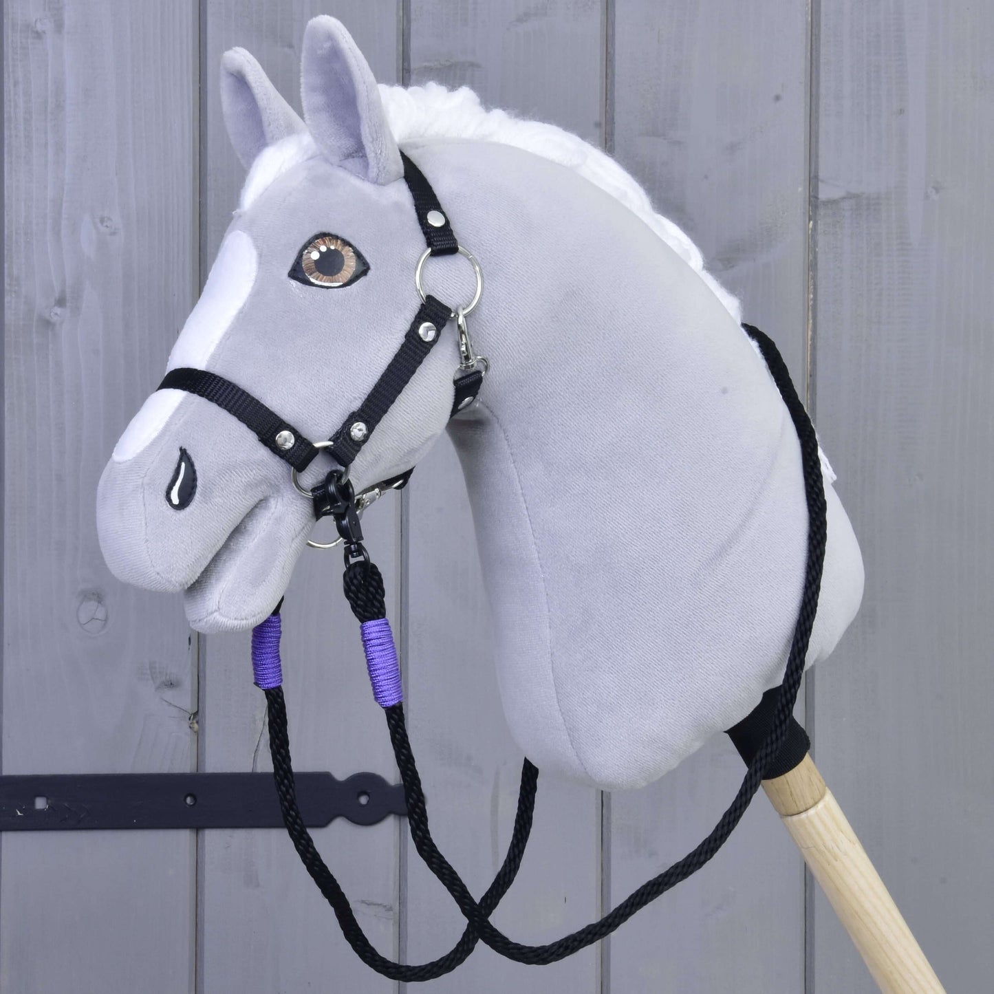 Hobby Horse LarDen Zügel Basic Lila, handgefertigt aus Polypropylenseil, gezeigt mit einem grauen Hobby Horse aus Plüsch mit weißer Mähne und schwarzem Zaumzeug für Hobby Horse, auf einem Holzstab vor einem grauen Holzzaun.