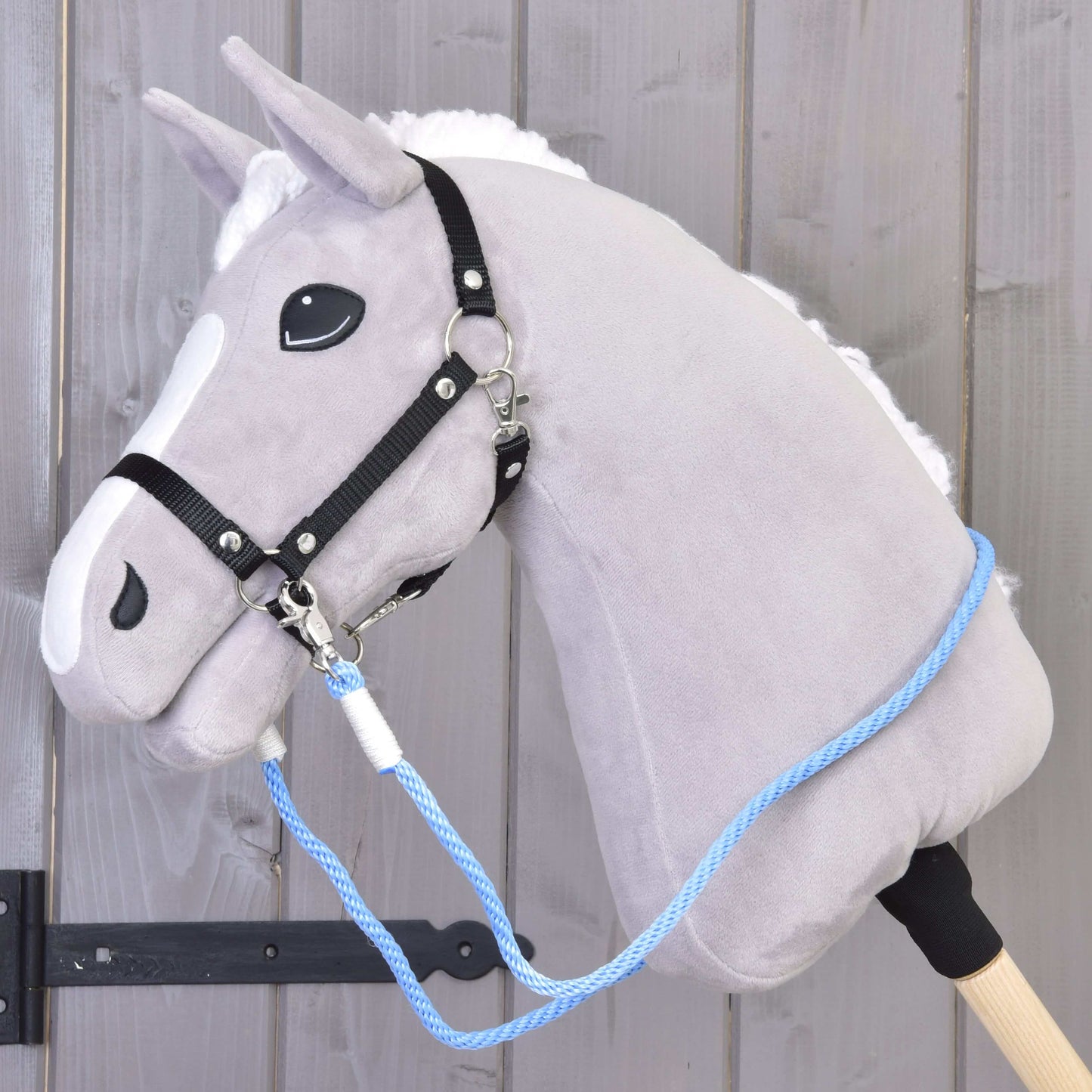 The Hobby Horse LarDen "Reins Sky" is a gray plush hobby horse with a black bridle, blue polypropylene rope, and handmade reins on a wooden stick, shown in front of a wooden wall.