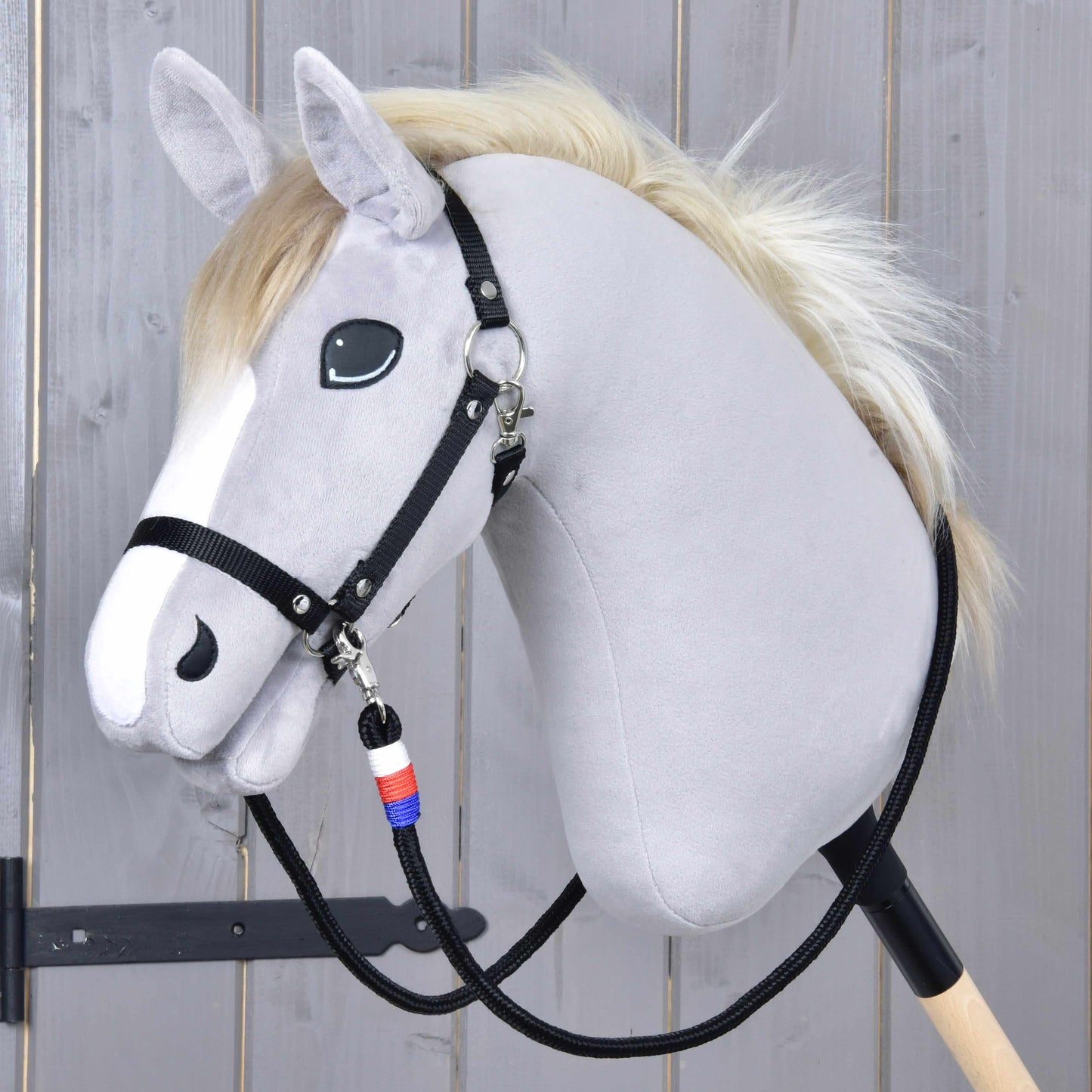 Rênes Emblème CZ de Hobby Horse LarDen avec Hobby Horse en peluche au visage gris clair, bride noire et crinière blanche, monté sur un bâton en bois, avec rênes faites main ornées d'un pendentif bleu, blanc et rouge.