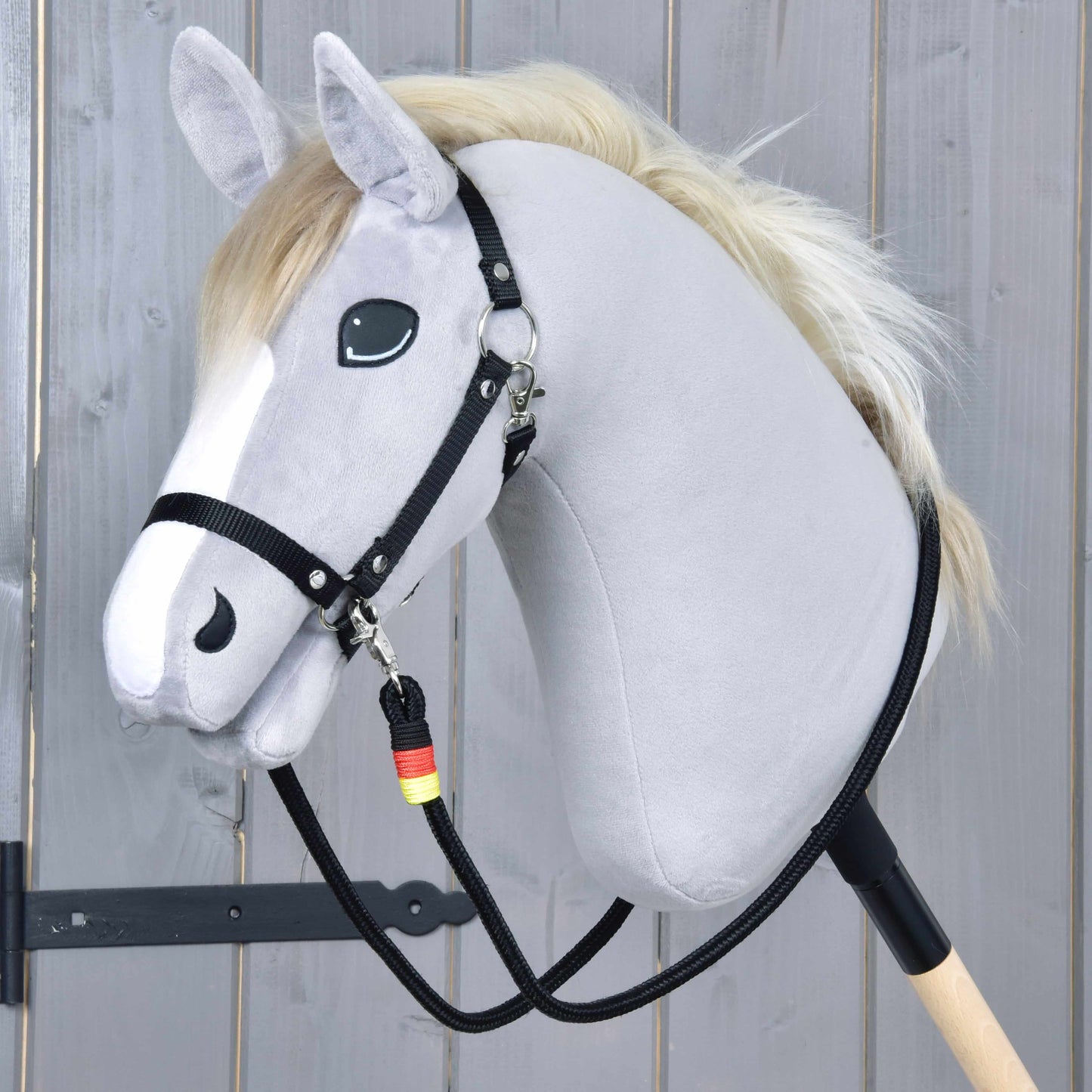 The Hobby Horse LarDen "Reins Emblem" is a gray plush hobby horse with a white mane, black bridle, and handmade polypropylene reins on a wooden stick, shown against a light wood plank background.
