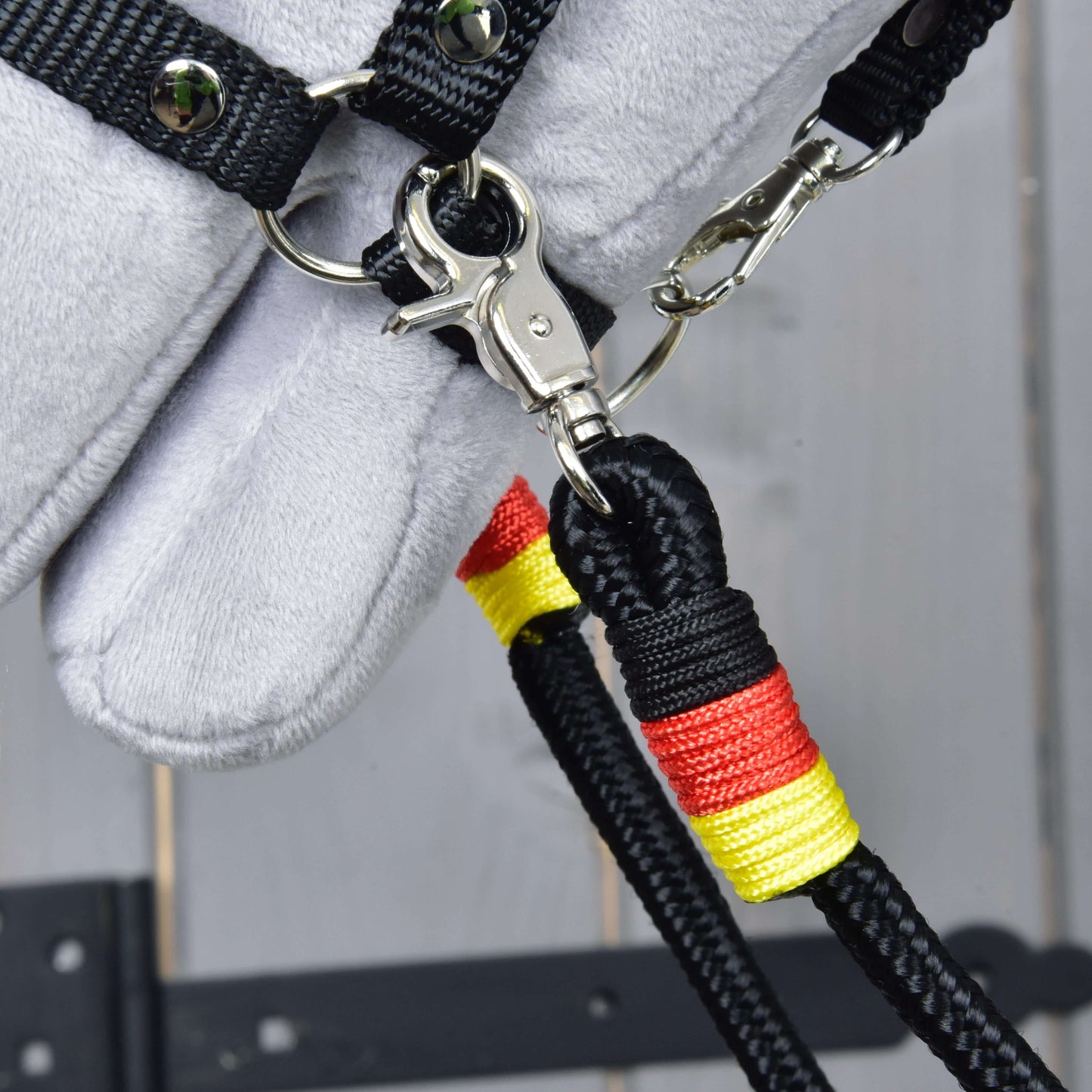 Close-up of Hobby Horse LarDen's Reins Emblem – black polypropylene rope reins with red and yellow wrapping, clipped to a metal ring on a black strap, attached to a grey plush model horse head against a wooden background.