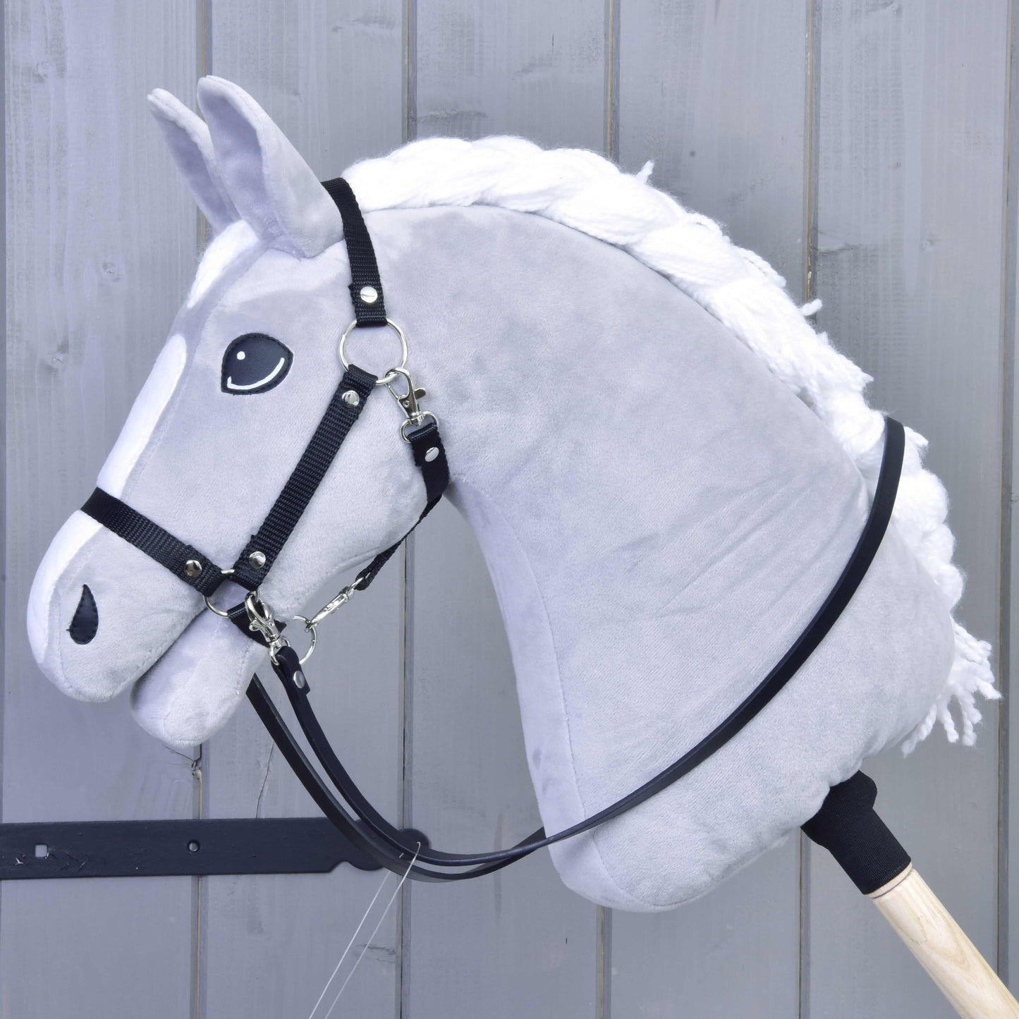 Zügel Eko Kunstleder von Hobby Horse LarDen an einem grauen plüschigen Hobby Horse mit weißer Mähne, gebisslosem Zaumzeug und schwarzen Kunstlederzügeln auf einem Holzstab, vor grauen Holzpaneelen.