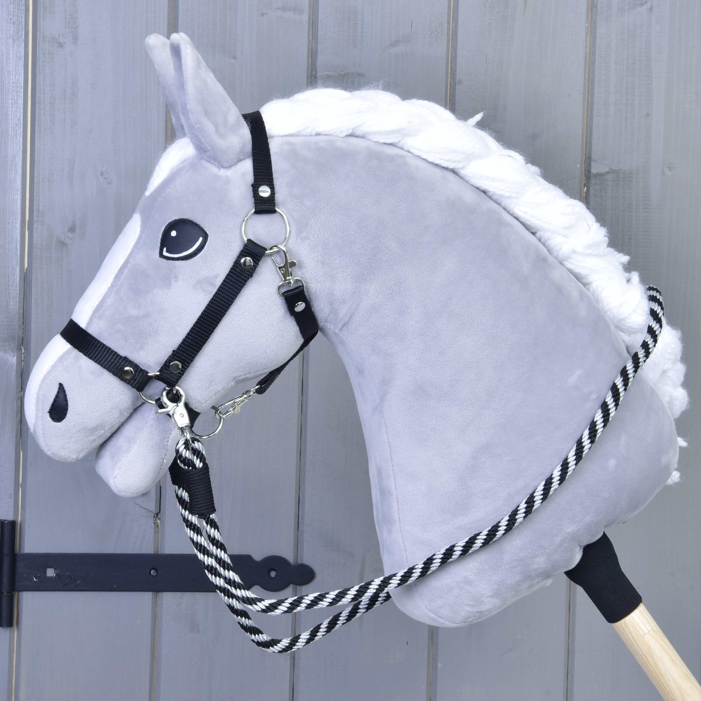 Hobby Horse - Rênes à rayures noires de Hobby Horse LarDen sur un Hobby Horse gris en peluche avec crinière blanche et bride noire, fait main, avec des rênes rayées noir et blanc en polypropylène, monté sur un bâton en bois devant un fond en bois gris clair.