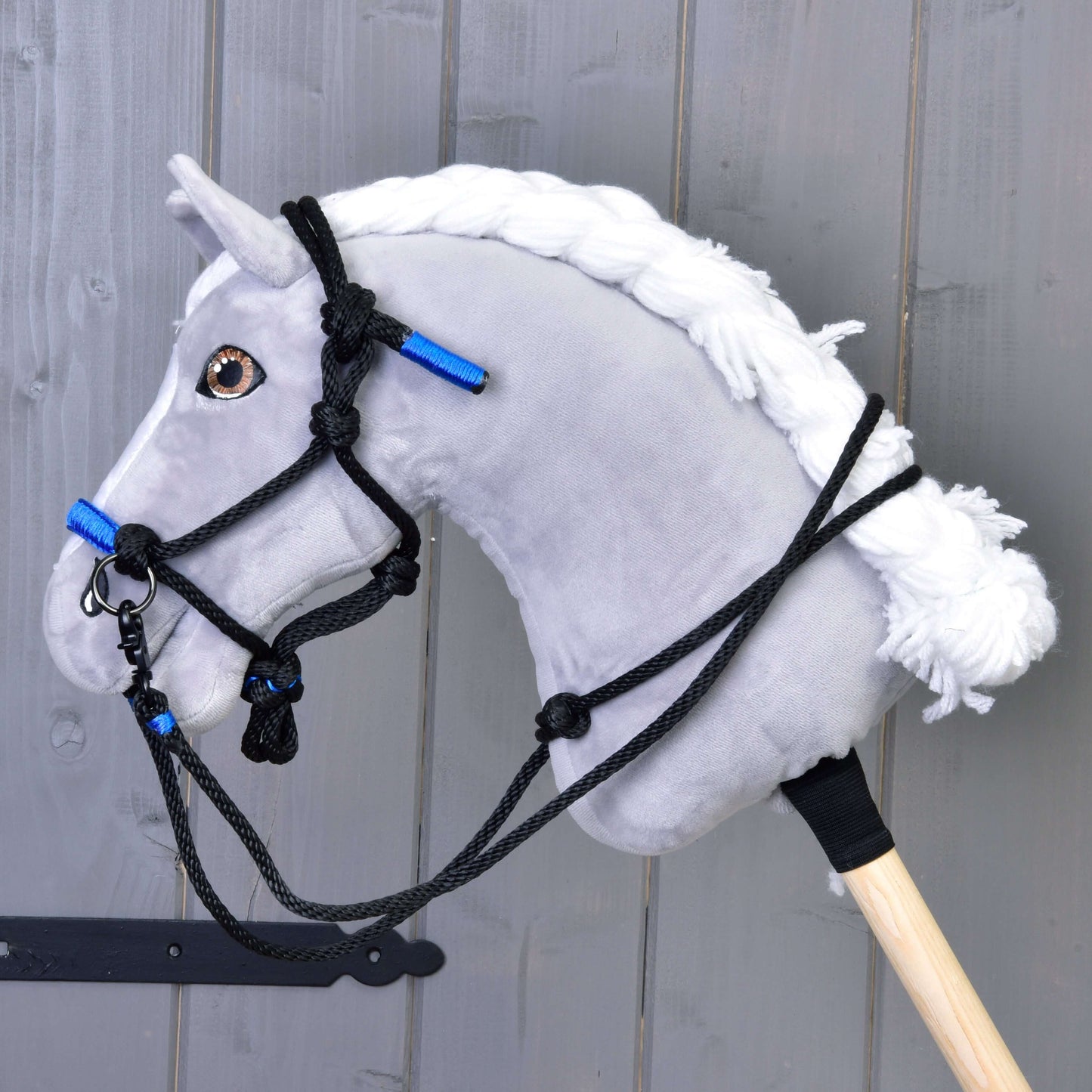 Seilhalfter Azure von Hobby Horse LarDen an einem grauen Plüsch-Hobby Horse mit weißer Garnmähne, schwarzem Zaumzeug und markanten azurblauen Akzenten, montiert auf einem Holzstab vor grauer Holzwand.