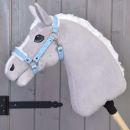 Ein graues Steckenpferd aus Plüsch mit weißer Mähne trägt das Halfter mit Fell - Kunstleder Pastell Blue von Hobby Horse LarDen und lehnt vor einer Holzwand mit Paneelen und schwarzen Metallscharnieren.