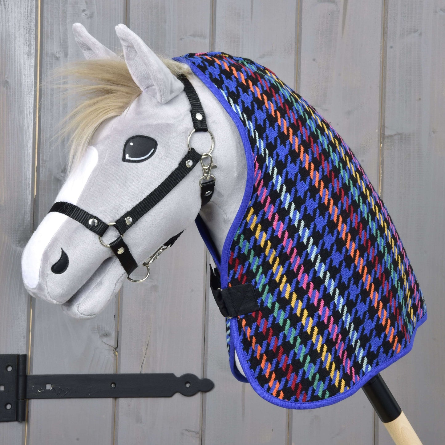 Un Hobby Horse blanc avec un licou noir porte la couverture d'écurie Pérou colorée de Hobby Horse LarDen, avec un motif pied-de-poule, sur fond en bois clair.
