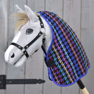 A white toy hobby horse with a black halter wears the colorful Stable Blanket Peru by Hobby Horse LarDen, featuring a houndstooth pattern, set against a light wooden background.