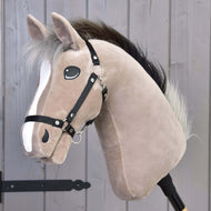 Hobby Horse Cloony mit Schwarzem Halfter von Hobby Horse LarDen, hellbraunes Plüsch-Sportpferd vom Typ Holsteiner mit weißer Blesse, schwarzem Halfter, flauschiger Mähne und Holzstab, vor einer grauen Holzwand.