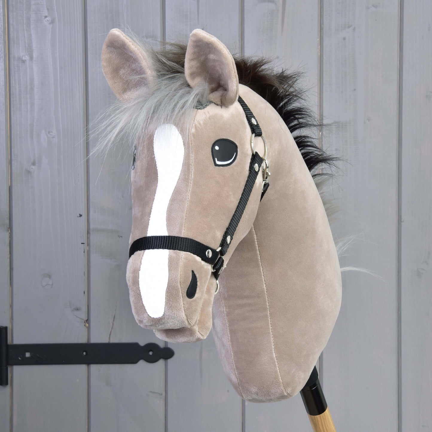 Hobby Horse Cloony mit Schwarzem Halfter von Hobby Horse LarDen, beiges Plüsch-Hobby Horse mit weißer Blesse, schwarzen Knopfaugen, schwarzer Mähne und Zaumzeug auf einem Holzstab – ideal als Spielzeug für Dressur.