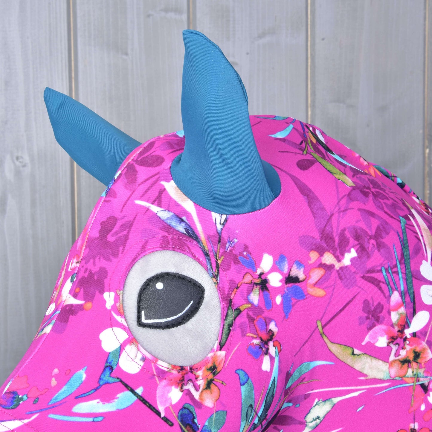 Combinaison de protection pour Hobby Horse Rose Beauté (Lycra) de Hobby Horse LarDen, avec tissu rose à motif floral, oreilles/cornes bleues en Lycra et grand œil brodé gris et noir, présentée sur fond en bois clair.