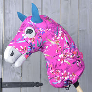 Hobby Horse LarDen „Hobby Horse Schutzanzug Pink Beauty (Lycra)“ mit leuchtend pinkem Blumenstoff, blauen Ohren und weißer Schnauze, montiert auf einem Holzstab vor einer grauen Holzwand.