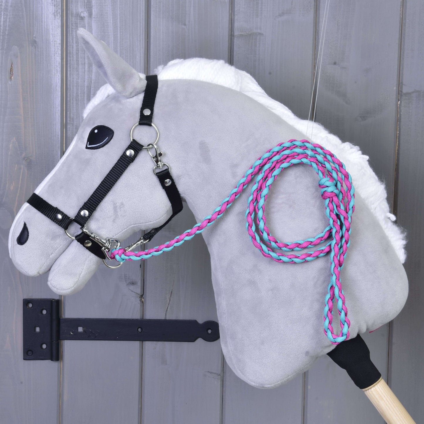 Hobby Horse LarDen Hobby Horse - Führstrick Knitted Pinkie-Blue mit grauem Plüschkörper, schwarzem Zaumzeug, weißer Mähne und geflochtenem Führstrick in Rosa und Türkis, hängt an einer Holzwand. Stabgriff inklusive.