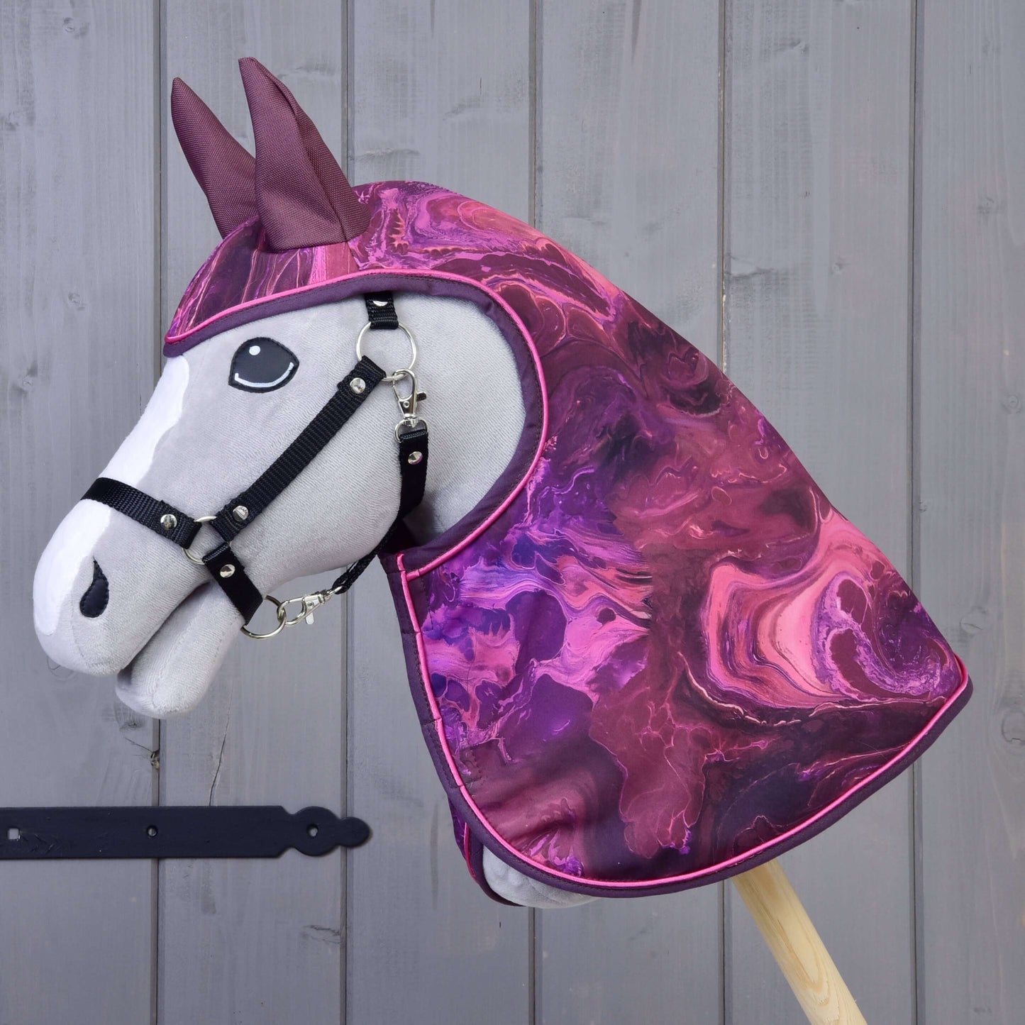 The Hobby Horse LarDen Raincoat Galaxy Purple features a plush hobby horse with gray face, black bridle, and a purple-pink marbled raincoat with matching ears—ideal for all-weather riding fun, shown against a wooden background.
