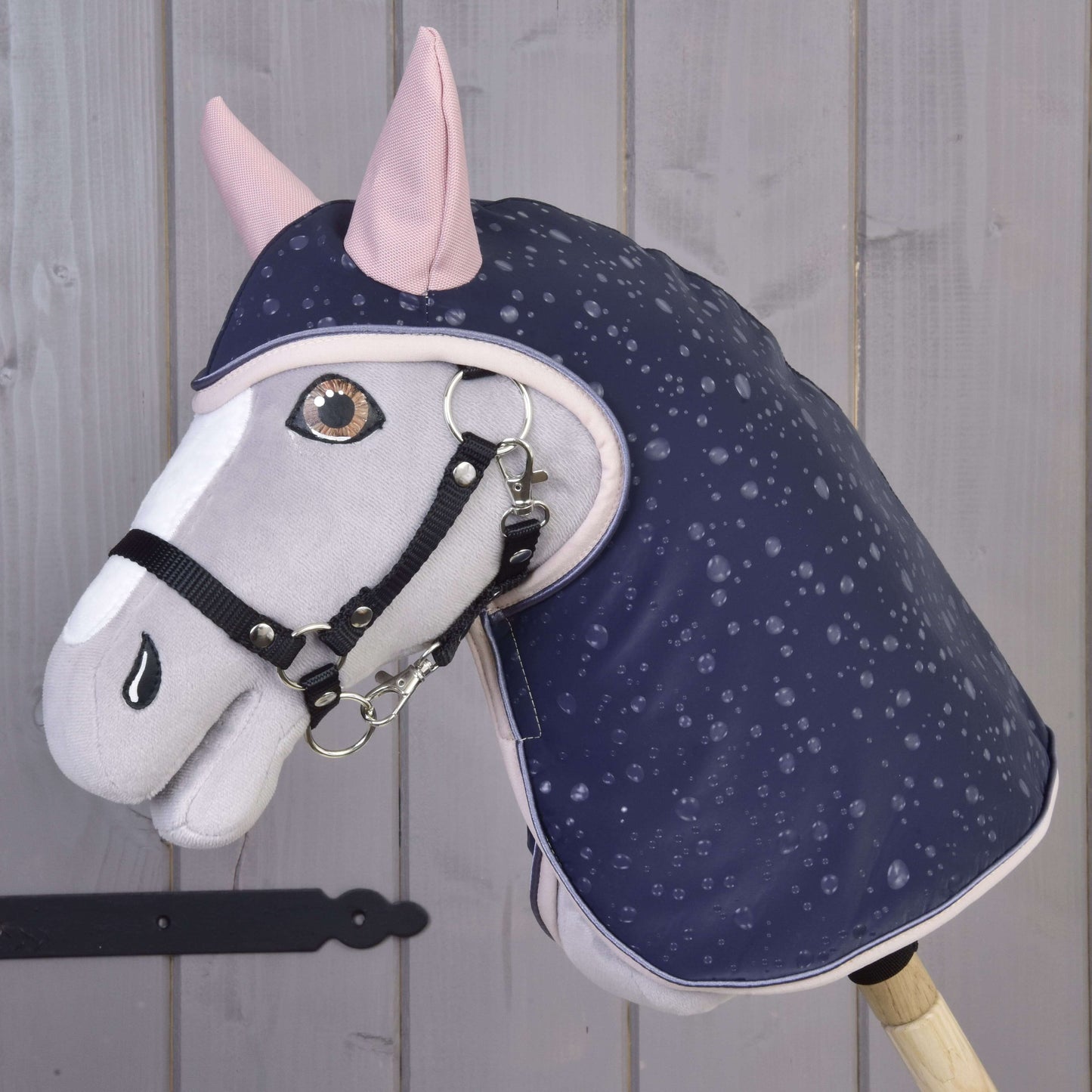 Die Hobby Horse LarDen Regendecke Pinkie Blue (softshell) zeigt ein grau-weißes Hobby Horse mit rosa Ohren und einer dunkelblauen Decke mit Regenmuster, schwarzem Zaumzeug, perfekt für Spiel bei jedem Wetter, vor heller Holzvertäfelung.