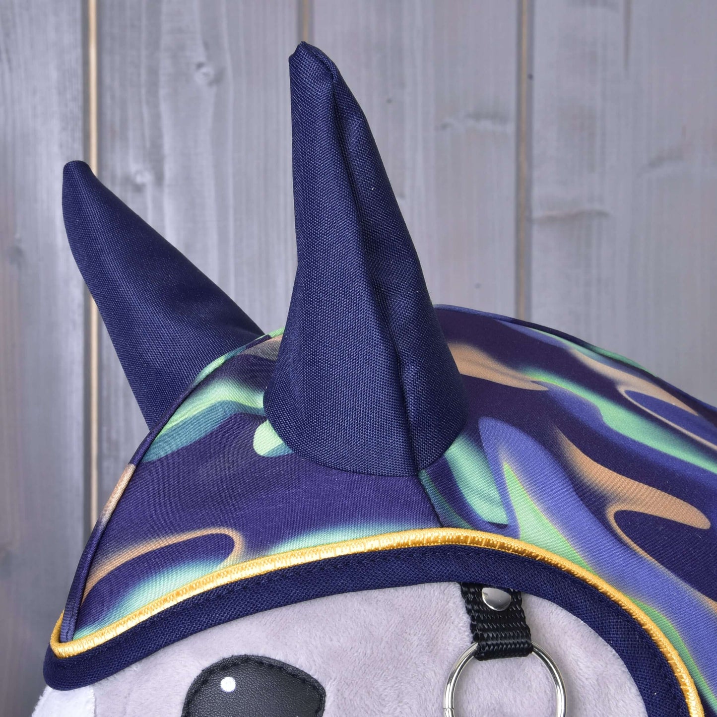 A close-up of Hobby Horse LarDen’s "Raincoat rainbow": a plush toy in a colorful tie-dye hood with two navy blue horns, styled like a whimsical waterproof raincoat, set against light gray wooden paneling.
