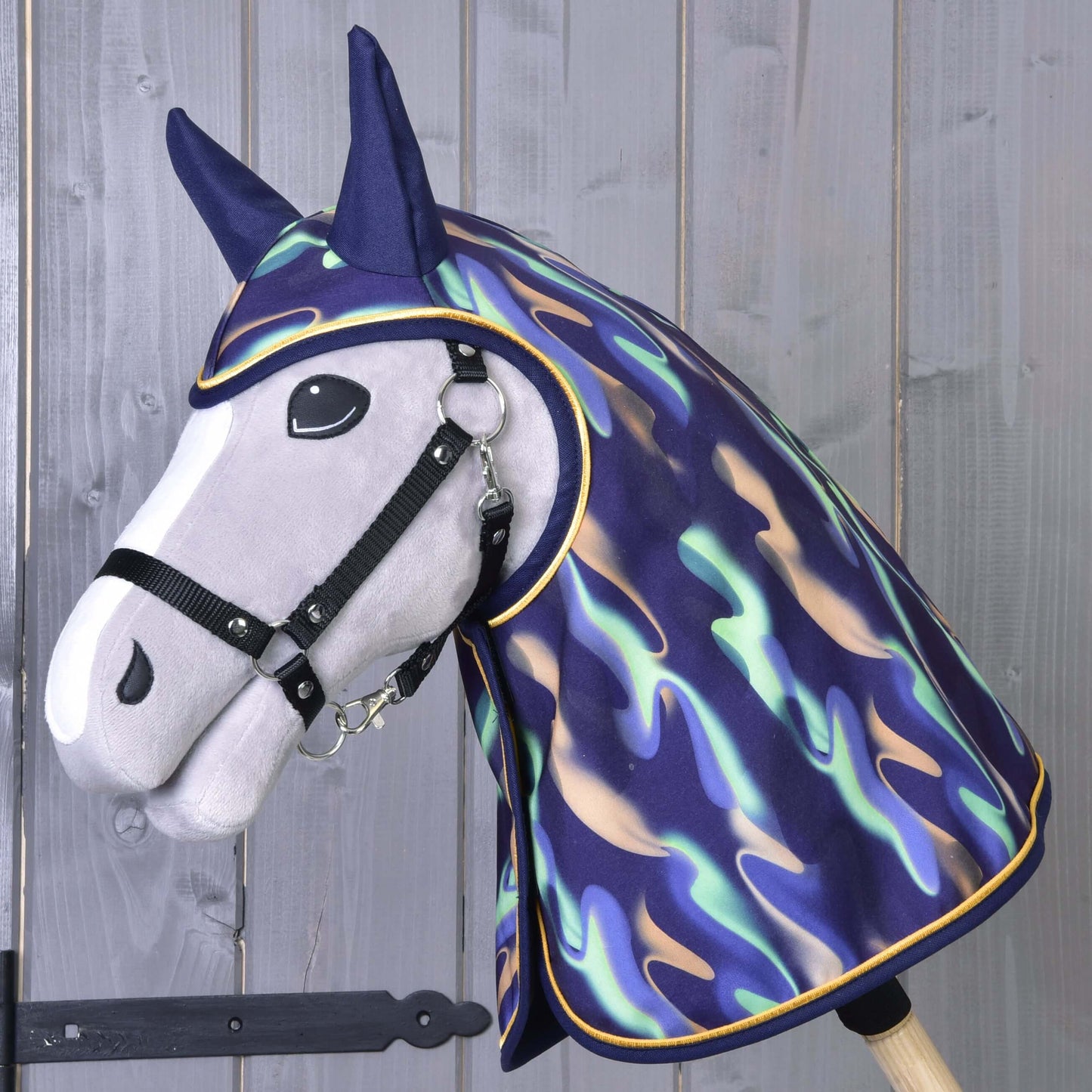 A plush hobby horse with a black bridle wears the Hobby Horse LarDen Raincoat rainbow, featuring a shiny multicolored design over its head and neck, set against a wooden background.