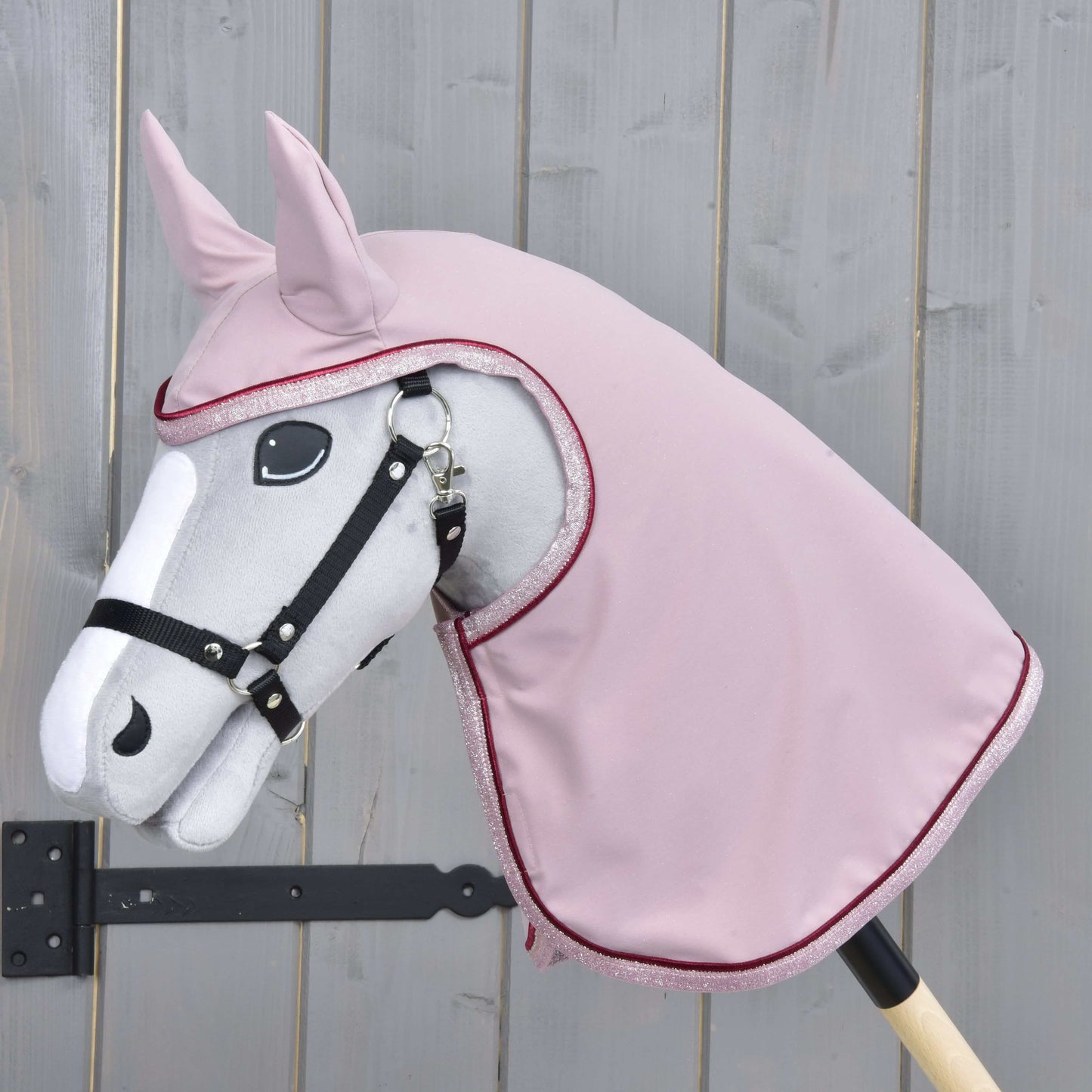 Das graue Hobby Horse LarDen auf einem Holzstab trägt ein schwarzes Zaumzeug und die Regendecke Veloria in Rosa über Kopf und Hals, vor dem Hintergrund eines hellen Holzzauns.