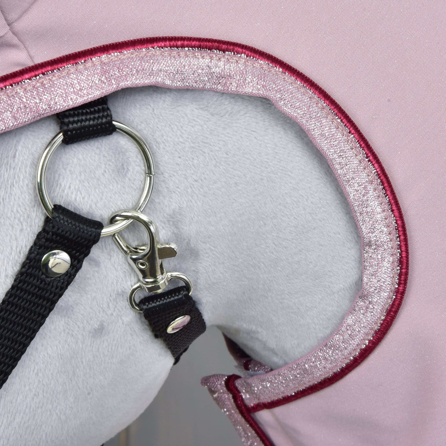 Nahaufnahme eines weißen Plüschtiers mit der Regendecke Veloria von Hobby Horse LarDen in Rosa mit glitzerndem Rand, mit schwarzer Leine an einem silbernen Ring und Karabiner.