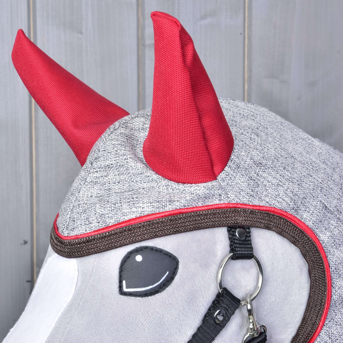 Eine Hobby Horse LarDen Regendecke Leonardo aus grauem Stoff mit leuchtend roten hornartigen Details und schwarzem Zaumzeug ist auf einem Pferd vor einem Holz Hintergrund zu sehen.