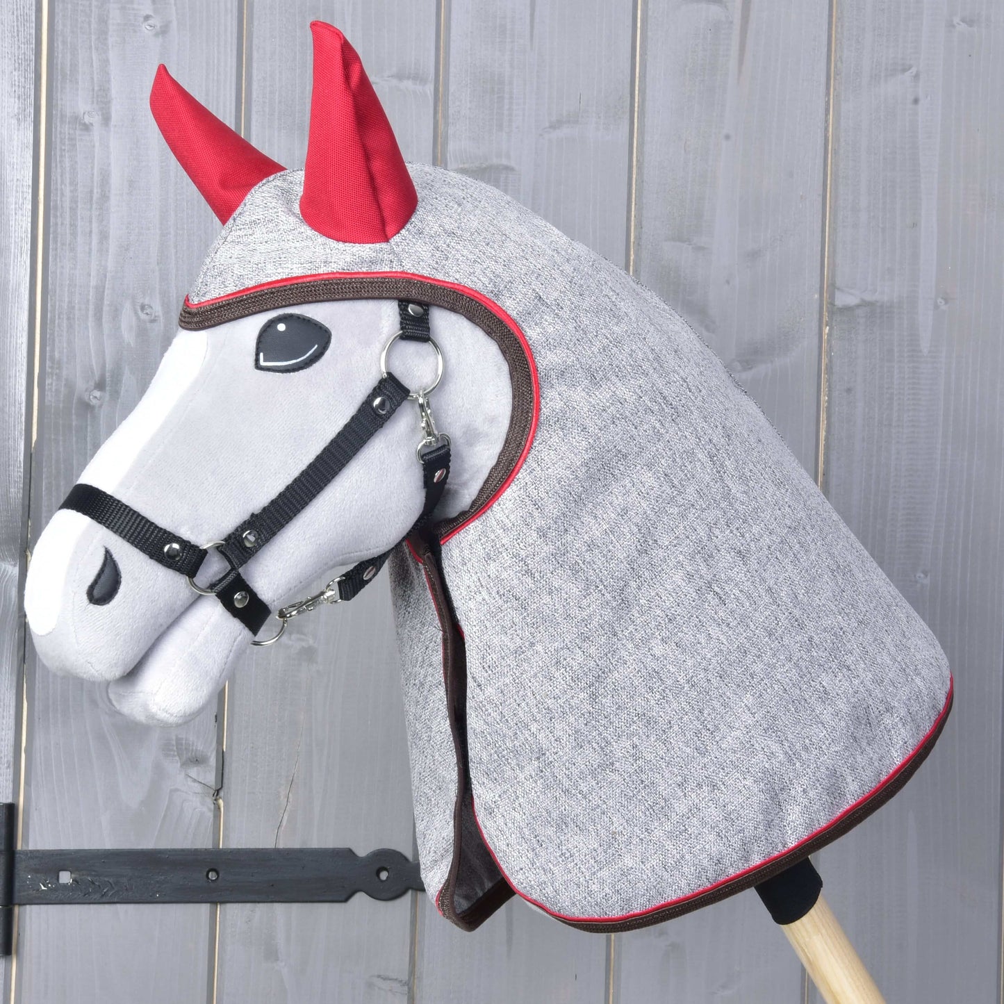 Die Regendecke Leonardo von Hobby Horse LarDen ist auf einem Plüsch-Hobby Horse mit grauer Kopfabdeckung, roten Ohren und schwarzem Zaumzeug auf einem Holzstab zu sehen – perfekt für Spiel bei jedem Wetter – vor einer grauen Holzwand.