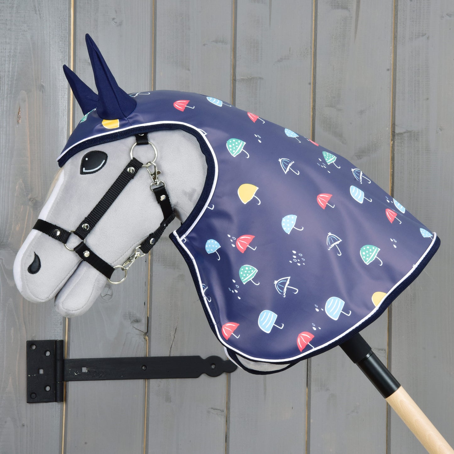 Hobby Horse LarDen "Raincoat Rainy", cheval en peluche avec bride et corne de licorne bleue, portant un imperméable bleu marine à parapluies colorés, monté sur un bâton en bois sur fond en bois gris — parfait pour jouer par tous les temps.