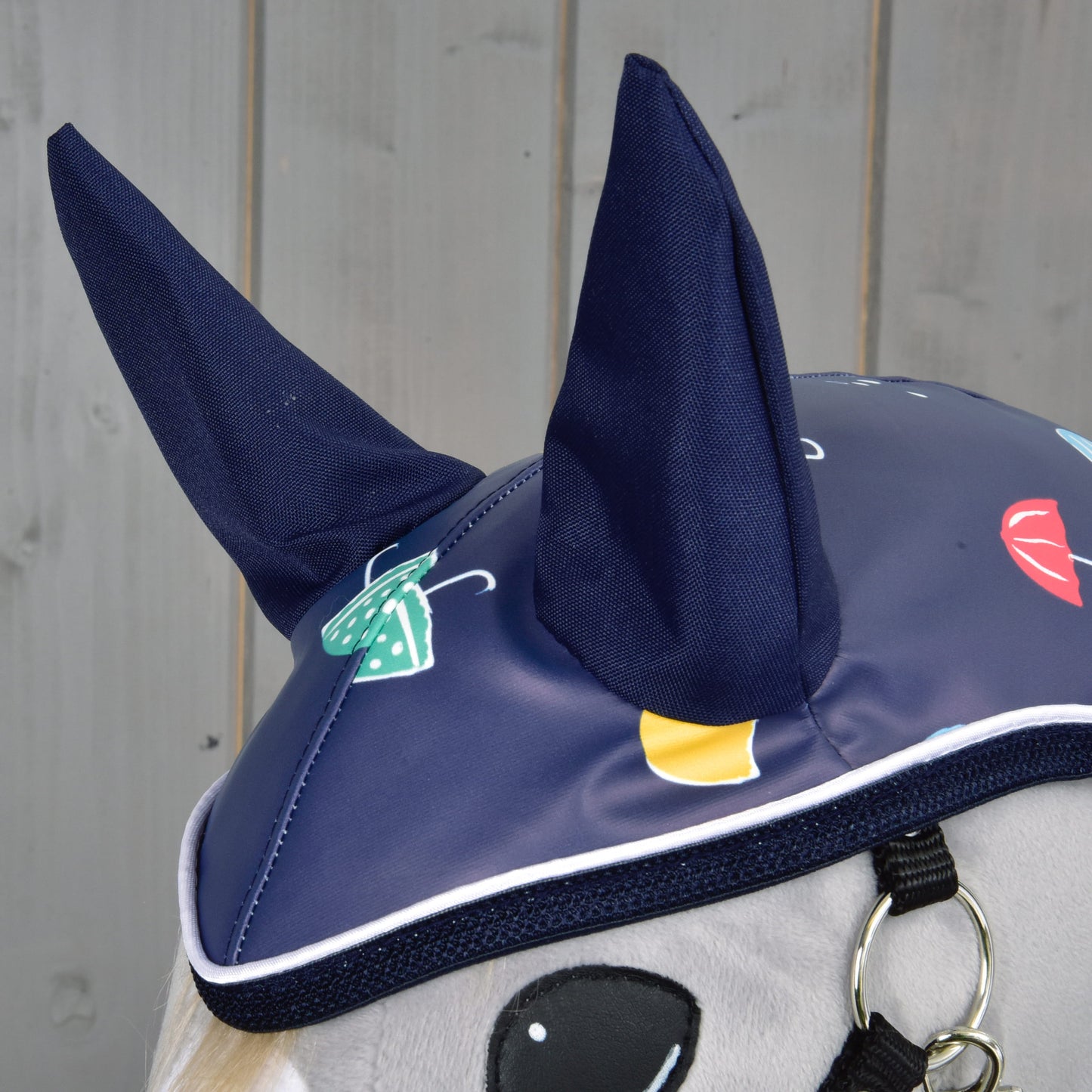 Gros plan sur le bonnet Imperméable Pluvieux bleu de Hobby Horse LarDen, avec oreilles dressées et imprimés vifs de parapluies et de gouttes de pluie — idéal par tous les temps. Sur fond de mur en bois gris clair.